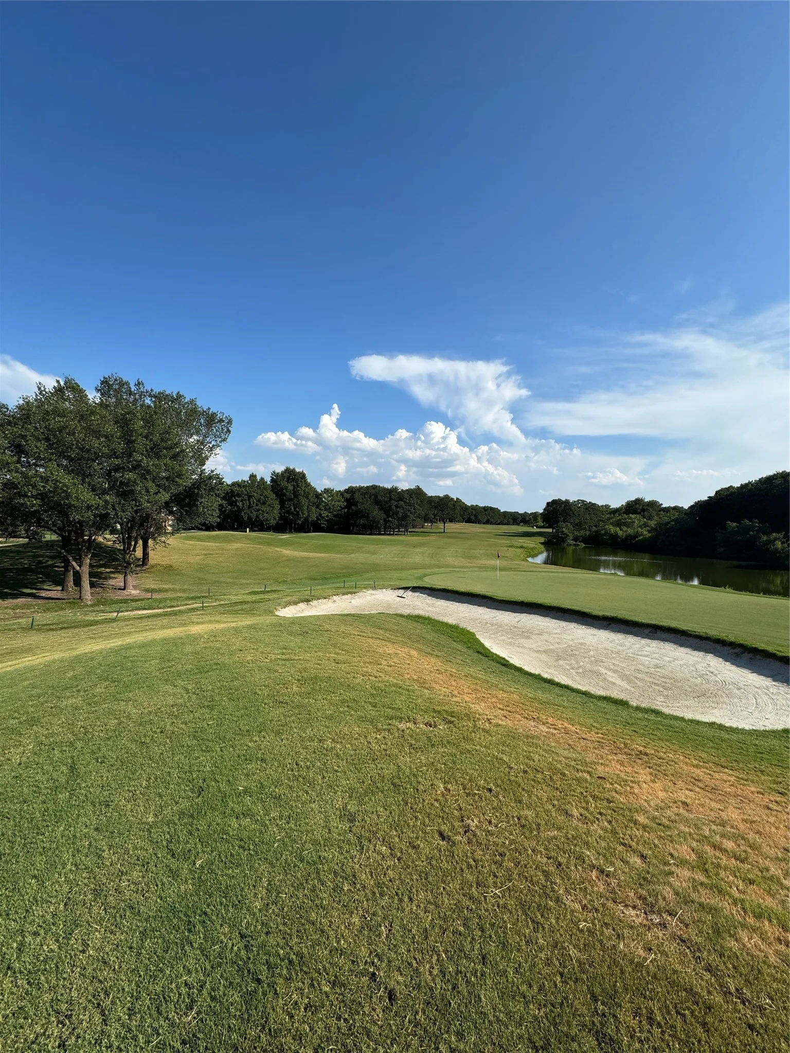 Golf course setting featuring expansive green lawns, a sand bunker, distant water features, and mature tree lines