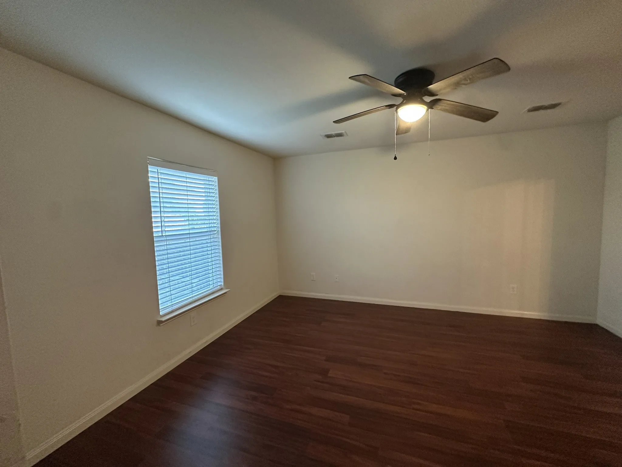 Spacious interior room featuring rich wood-finish flooring, a white ceiling fan with integrated lighting, and a window with blinds