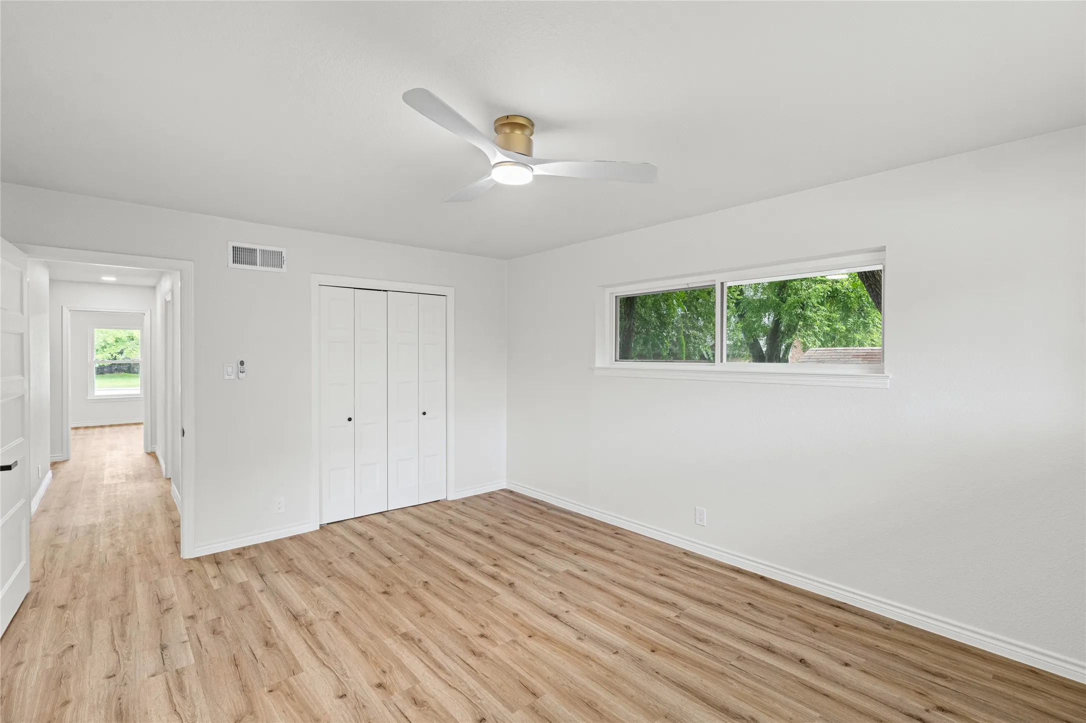 Room featuring wood-finish flooring, a contemporary ceiling fan with integrated lighting, and a horizontal window offering exterior tree views