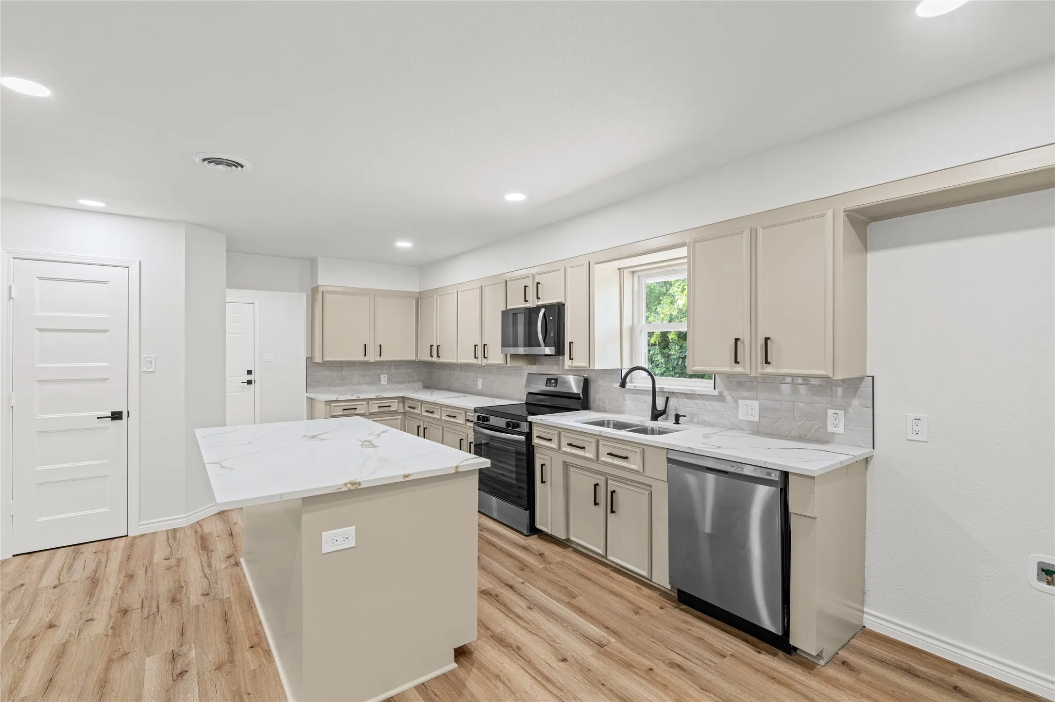 Modern kitchen featuring a central island with a stone-finish countertop, light-toned cabinetry, stainless steel appliances, and wood-finish flooring
