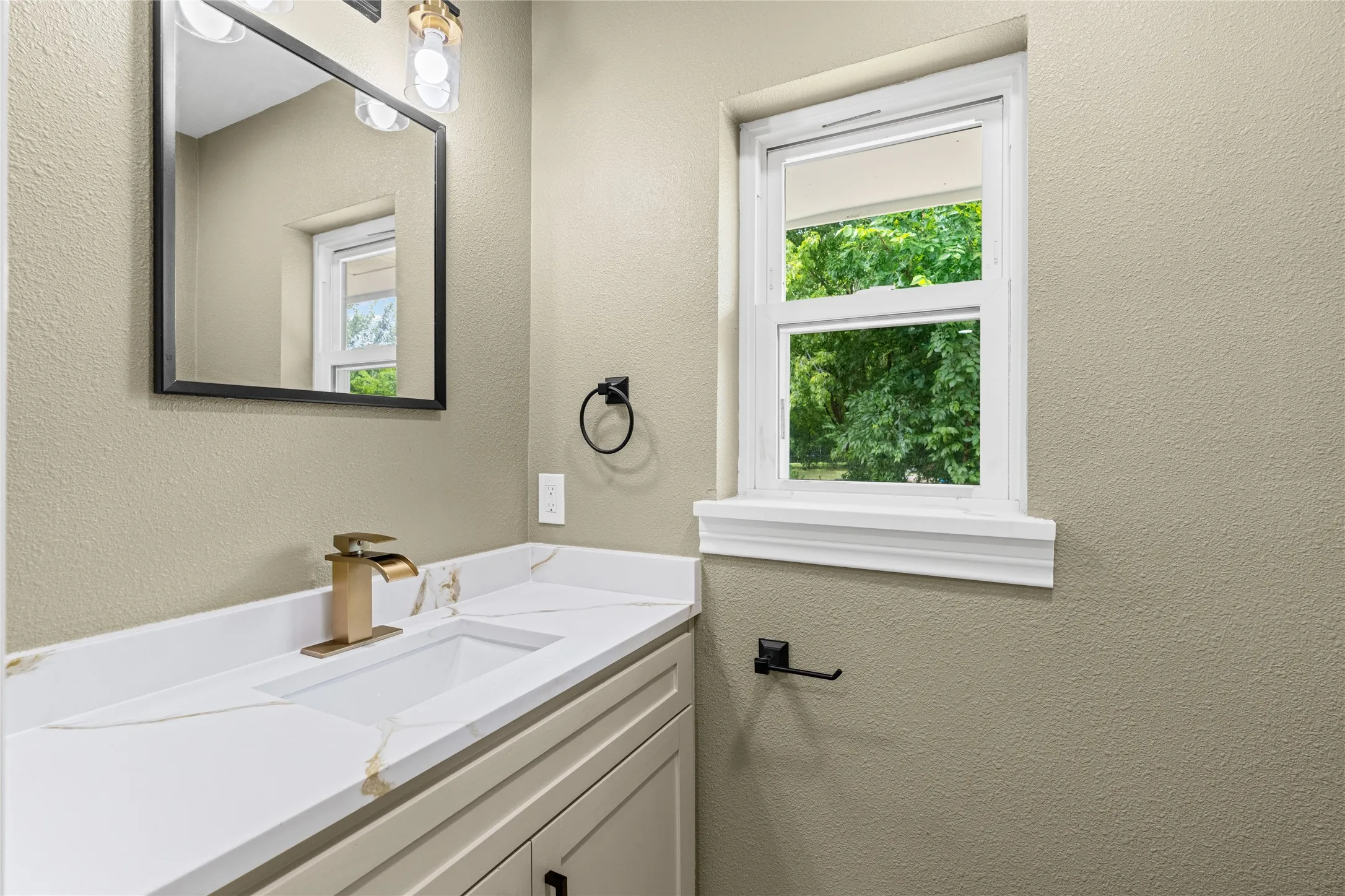 Bathroom featuring a single vanity with a white countertop and integrated sink, gold-finish faucet, and coordinating black-framed mirror