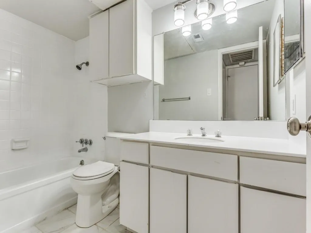 Bathroom featuring a white vanity with integrated sink, a large mirror, overhead lighting, a tub-shower combination with white tile surround, and built-in white cabinetry
