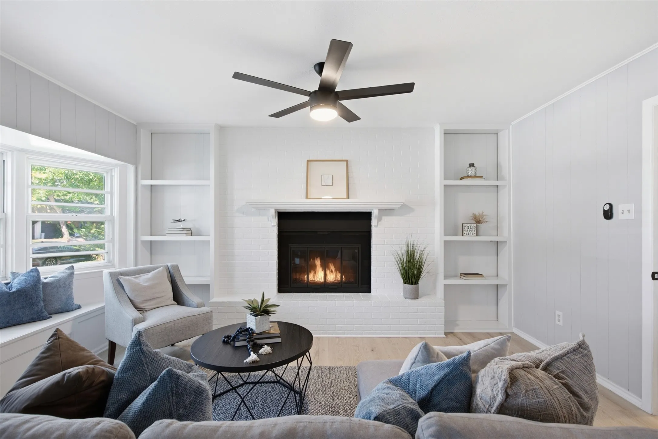 Gather Around the Fireplace — The converted garage now serves as a spacious living area anchored by a painted brick fireplace, built-in shelving, and a modern ceiling fan.