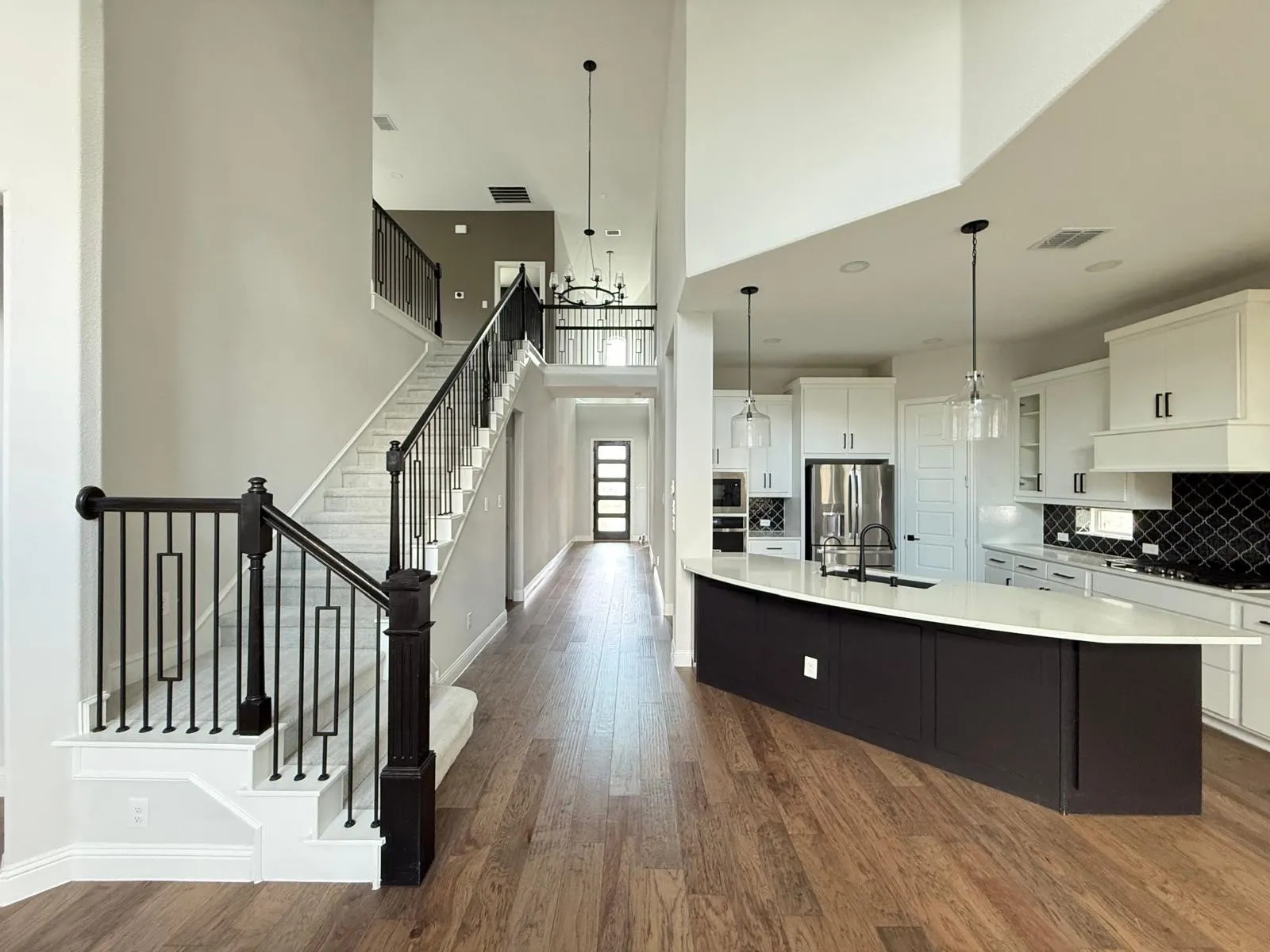Open concept interior featuring wood-finish flooring, a contemporary staircase with black wrought iron balusters, a large kitchen island with a light-colored countertop, white cabinetry, and a black diamond-patterned backsplash