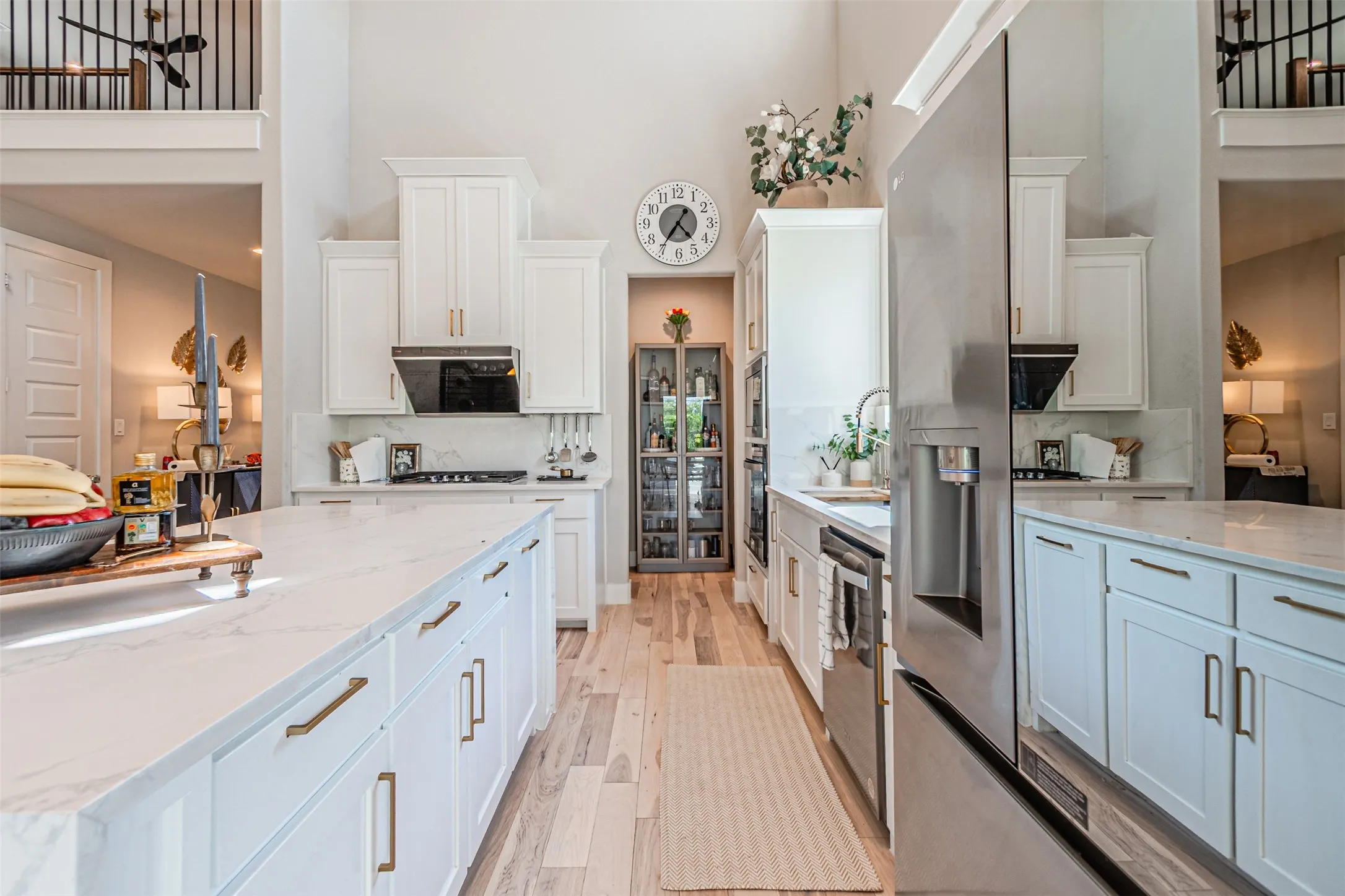 Spacious kitchen featuring an expansive island with a stone-finish countertop, high vaulted ceilings, light wood-finish flooring, white cabinetry with gold-tone hardware, and stainless steel appliances