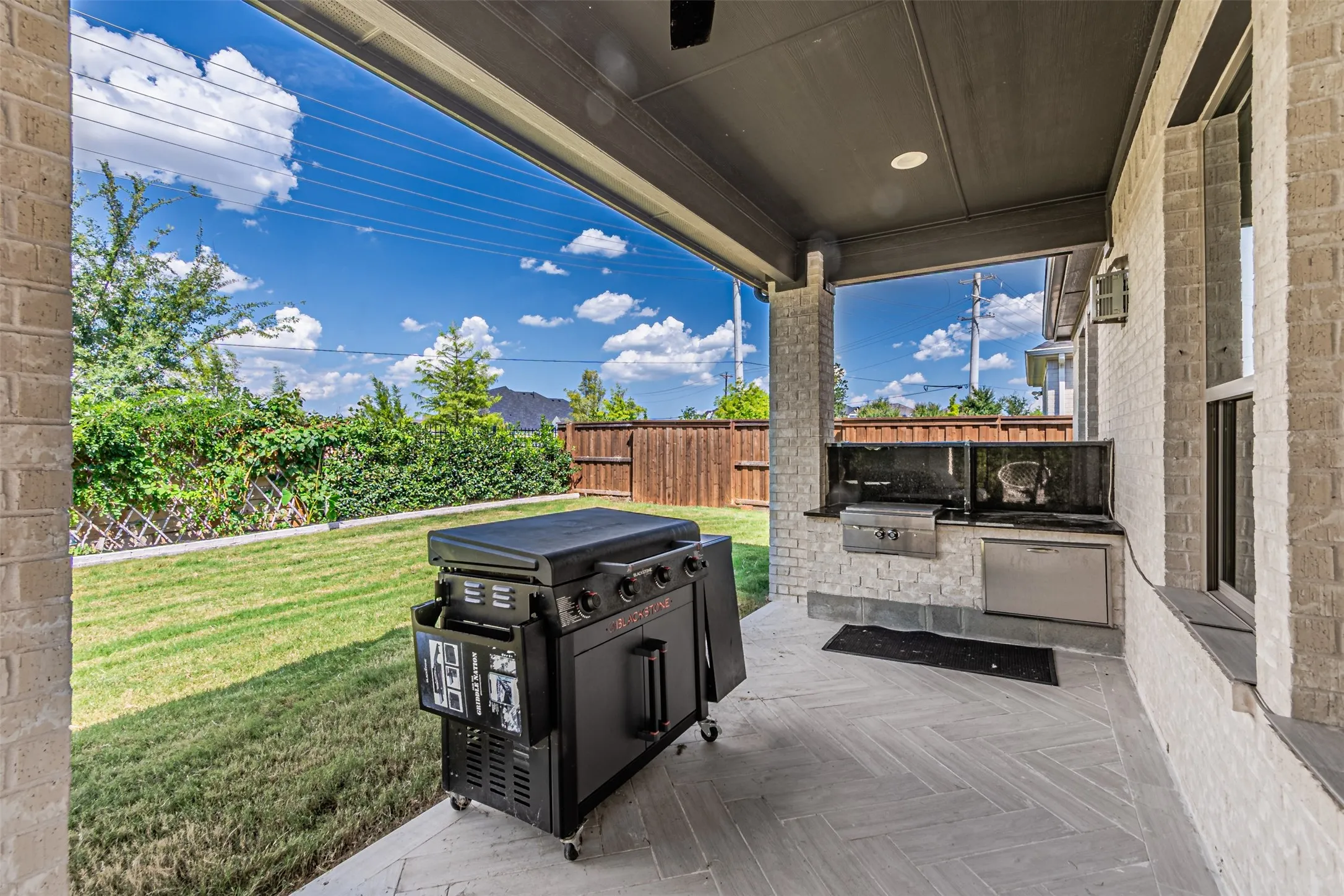 Covered outdoor patio featuring light brick columns, a dark ceiling with recessed lighting, and a built-in outdoor kitchen with a grill and stainless steel refrigerator