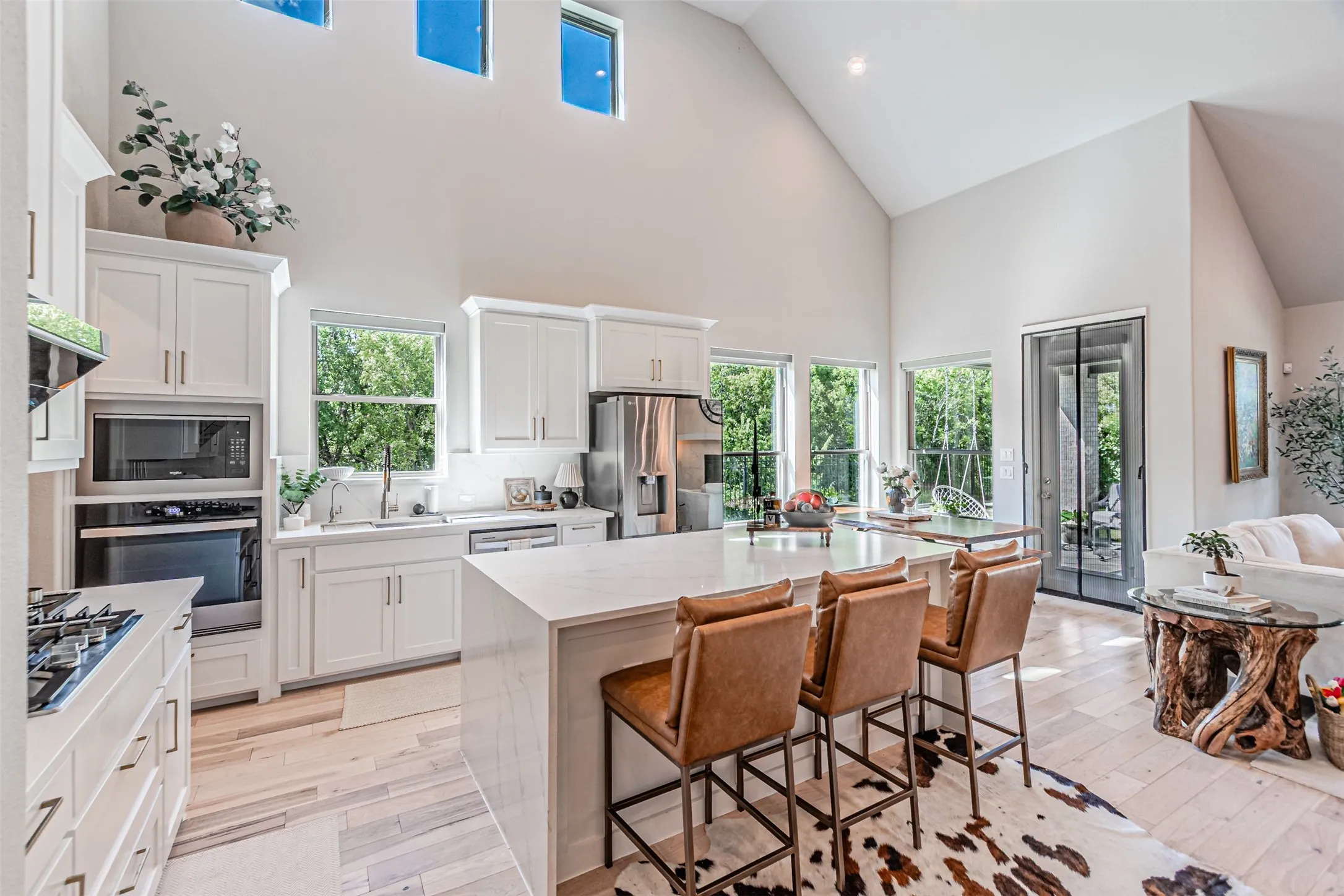 Vaulted ceilings featuring upper transom windows and a contemporary kitchen with white shaker cabinetry, stainless steel appliances, a center island with countertop seating, and light wood-finish flooring