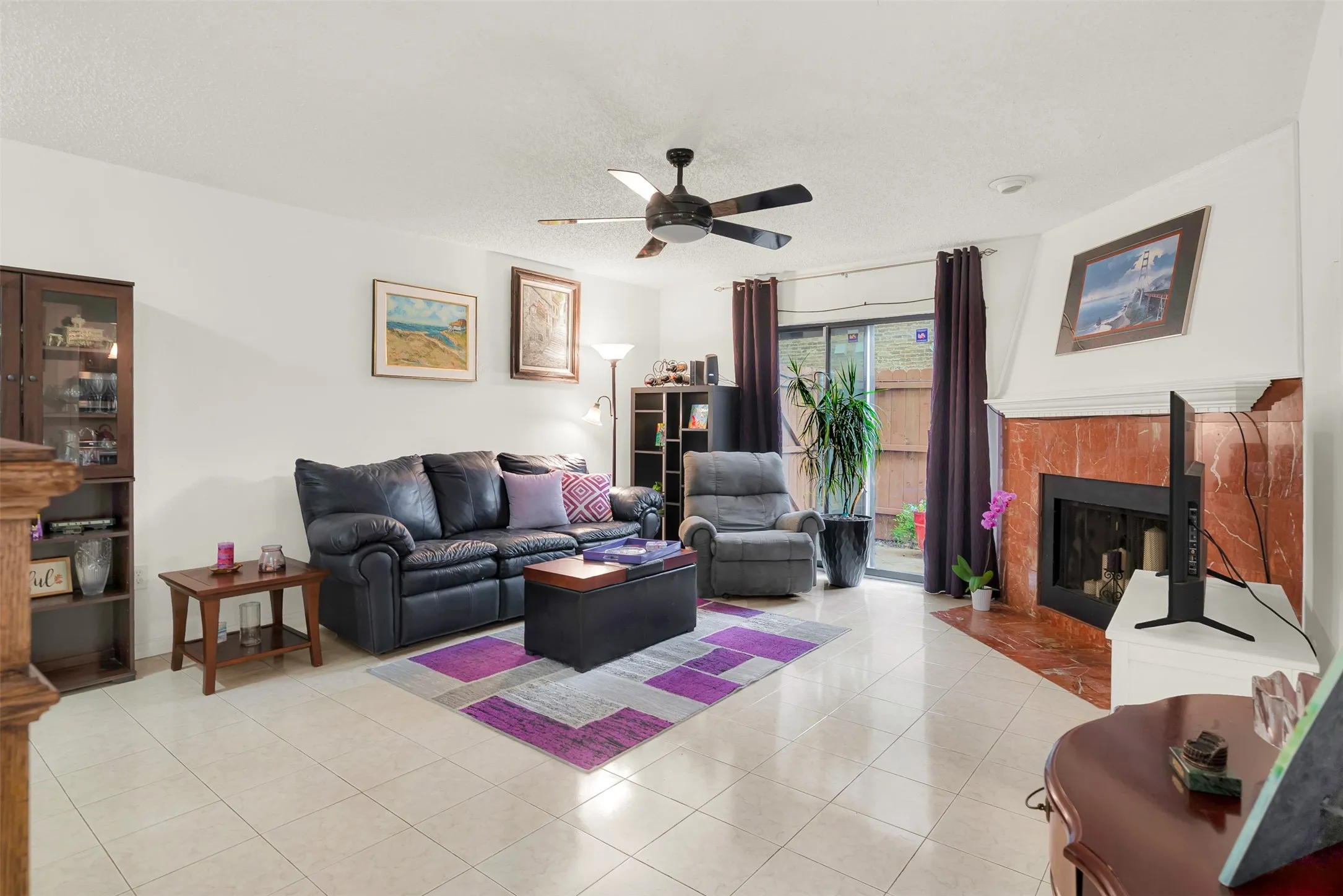 Living area featuring tile flooring, a fireplace with a stone surround, a ceiling fan, and sliding glass door to patio.