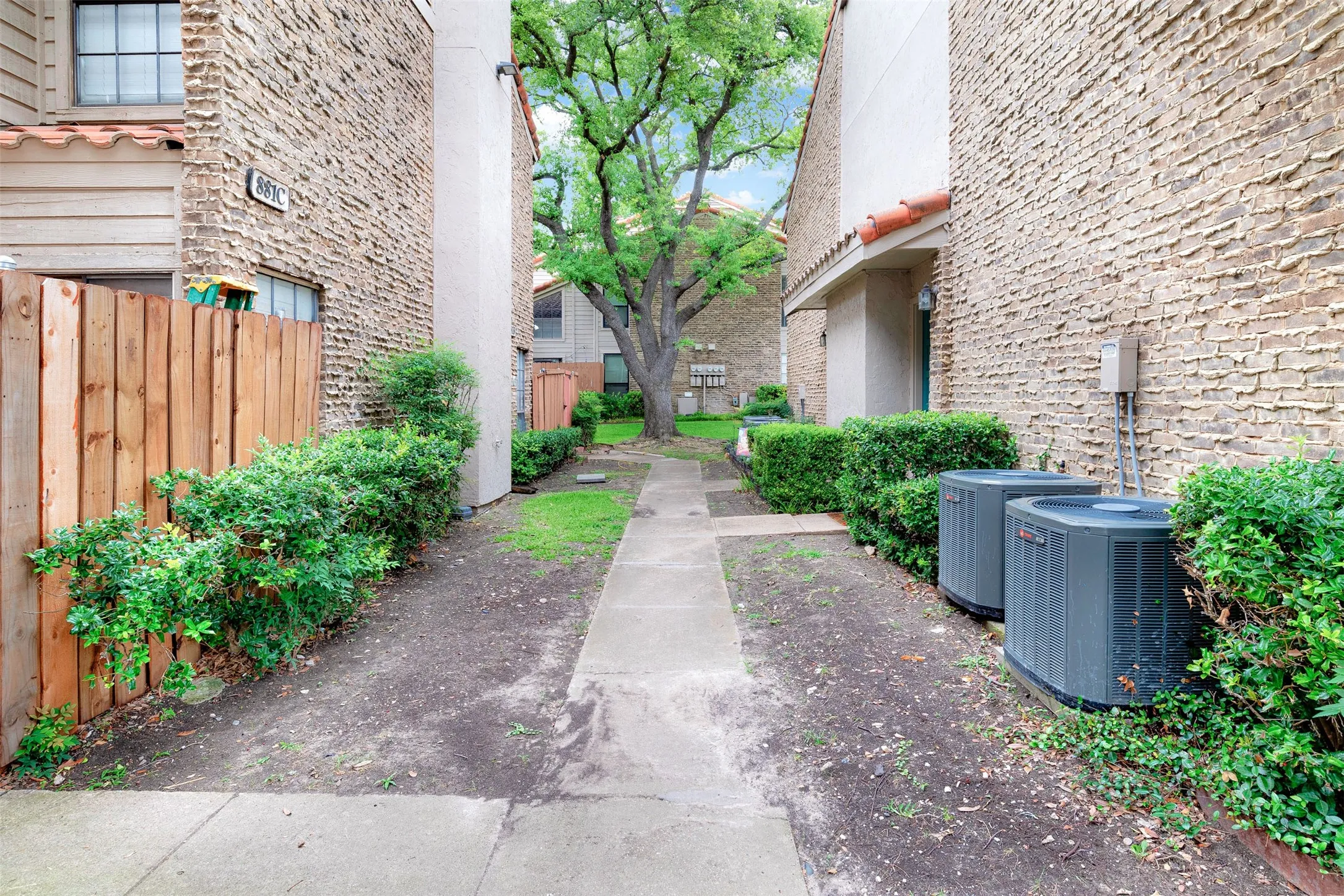 Pathway bordered by brick and stucco exteriors, featuring a privacy fence, mature landscaping, and central air conditioning units