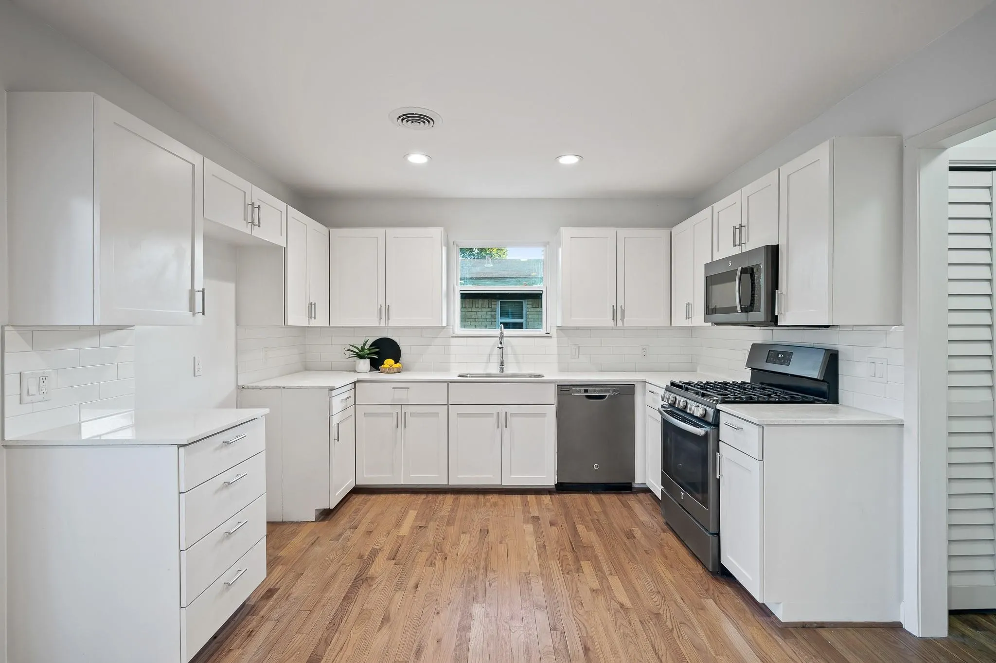 Open and updated kitchen featuring white shaker cabinetry, light-toned countertops, subway tile backsplash, and wood-finish flooring. Vacant older photo.
