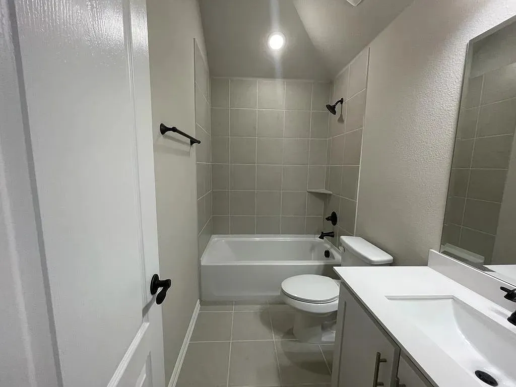 Bathroom featuring a single vanity with an integrated sink, a rectangular mirror, a toilet, a bathtub with a shower, and light-toned wall and floor tile