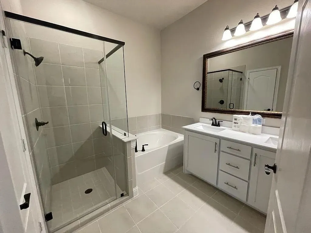 Bathroom featuring a walk-in shower with clear glass enclosure and black hardware, a built-in tub, dual vanity with white cabinetry and undermount sinks, and light-toned tile flooring