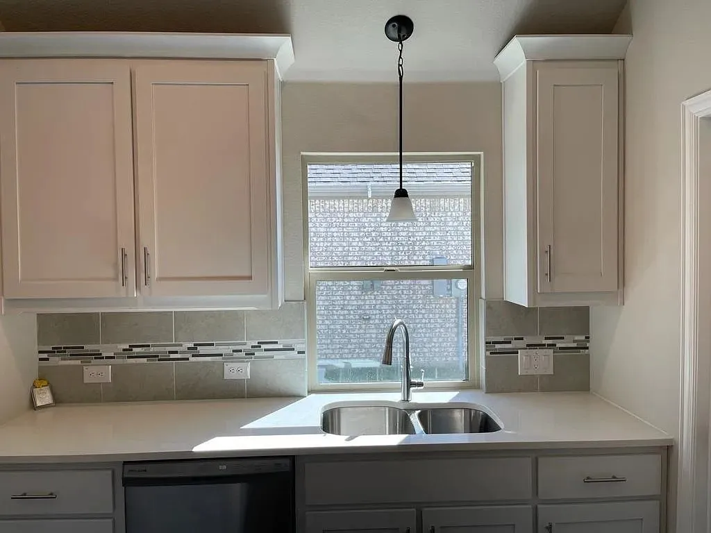 Kitchen sink area featuring light-toned cabinetry with crown molding, a stainless steel double basin sink with a gooseneck faucet, light-colored countertops, and a tile backsplash with a decorative mosaic accent