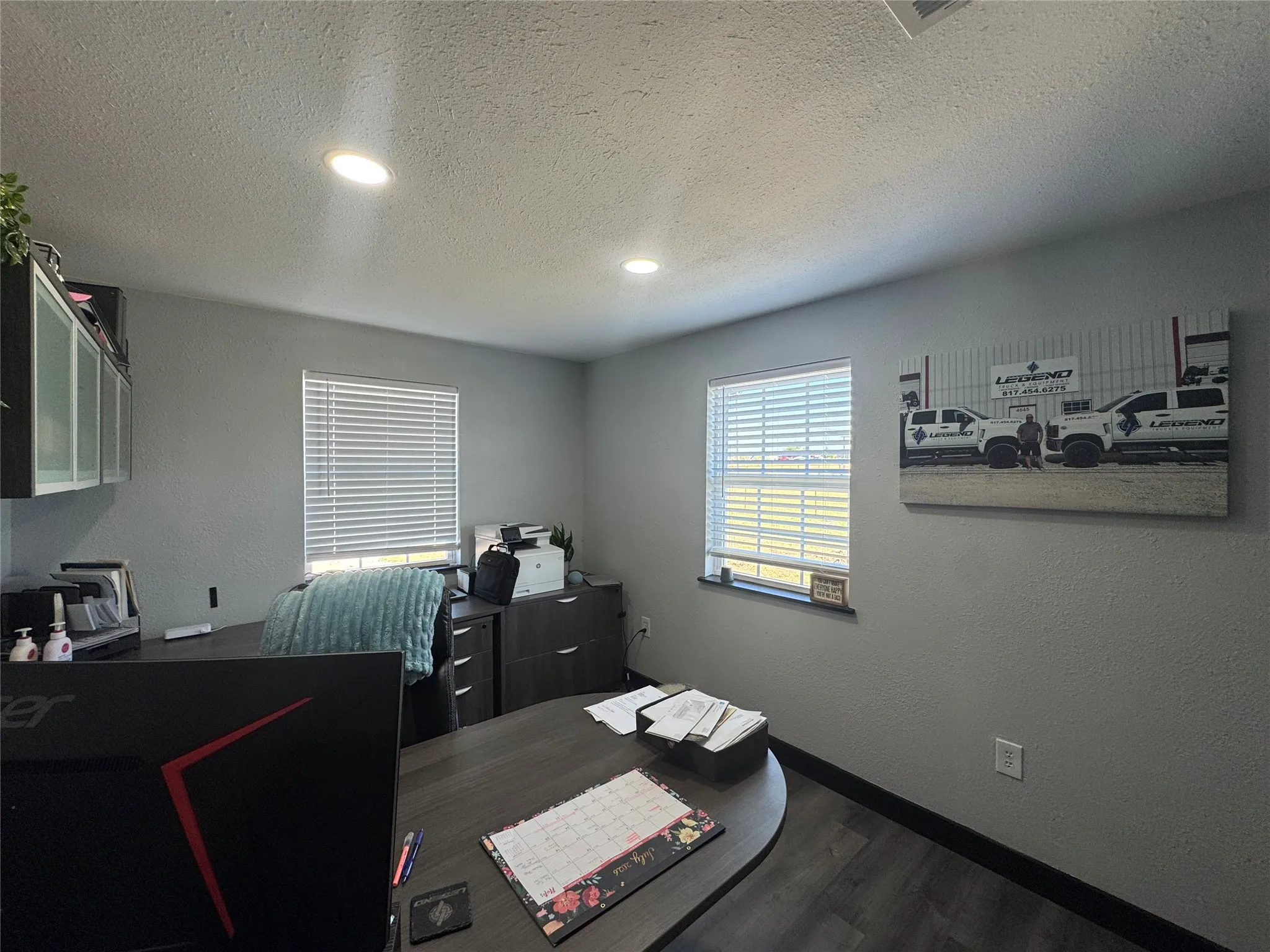 Office space featuring gray walls, wood-finish flooring, two windows with blinds, built-in upper cabinetry with frosted glass, and recessed ceiling lighting