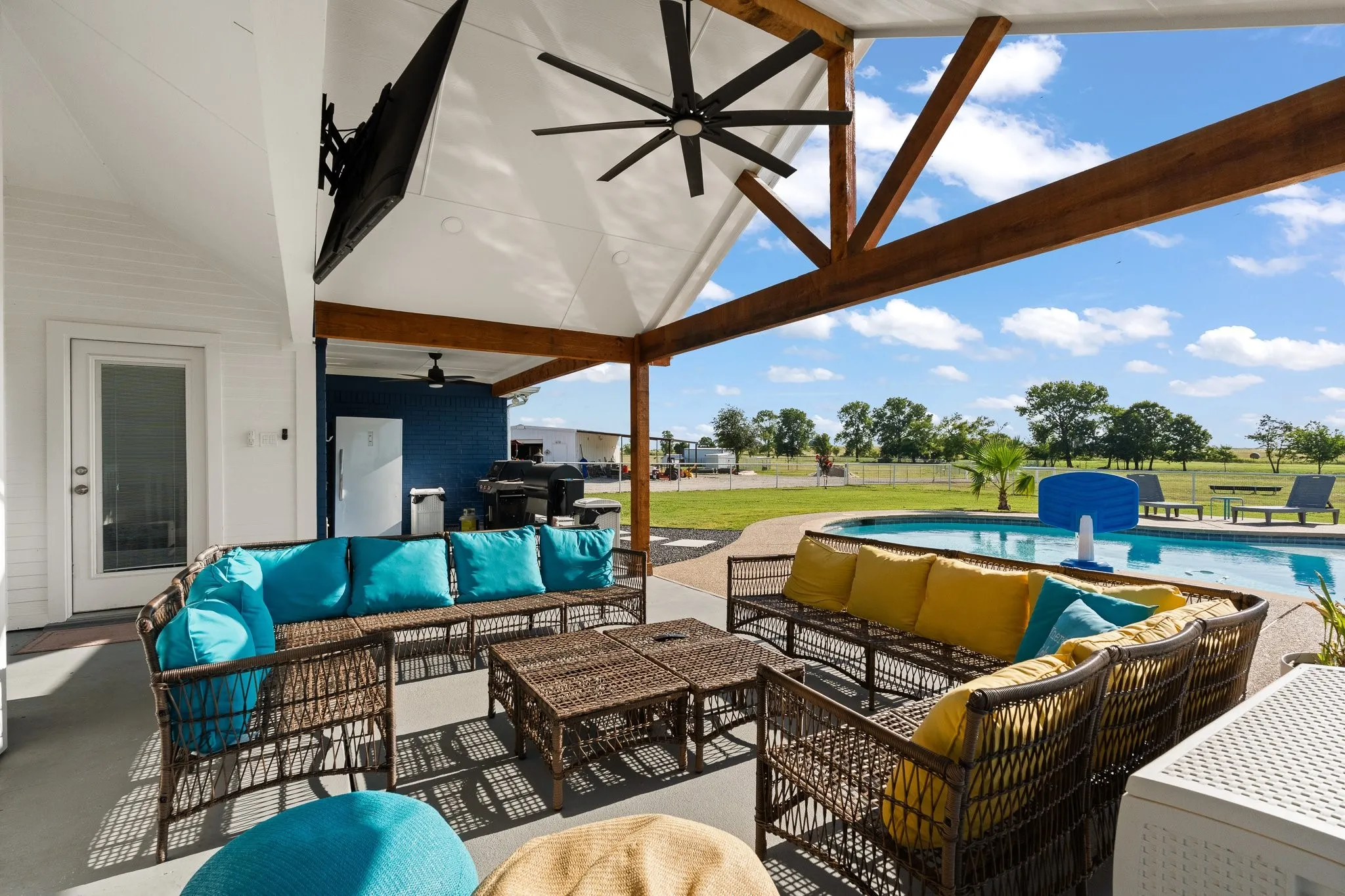 Covered patio with exposed wooden beams and a ceiling fan, featuring an outdoor television, an inground swimming pool, and a white exterior door