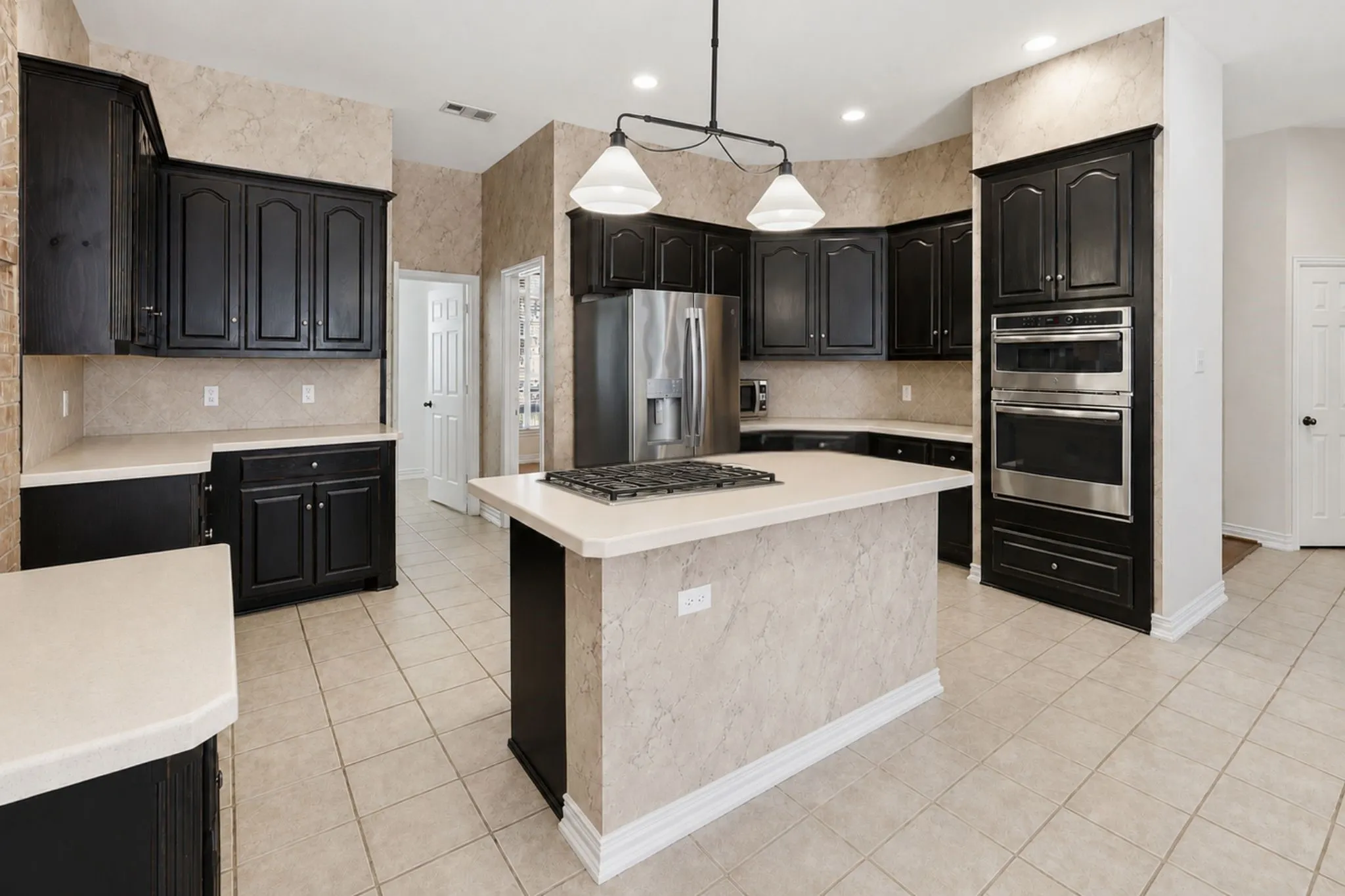 Spacious kitchen featuring dark wood cabinetry, a central island with an integrated cooktop, stainless steel appliances including a double oven and refrigerator, light-toned countertops, and tile flooring *Virtually Staged see other photos of the island*