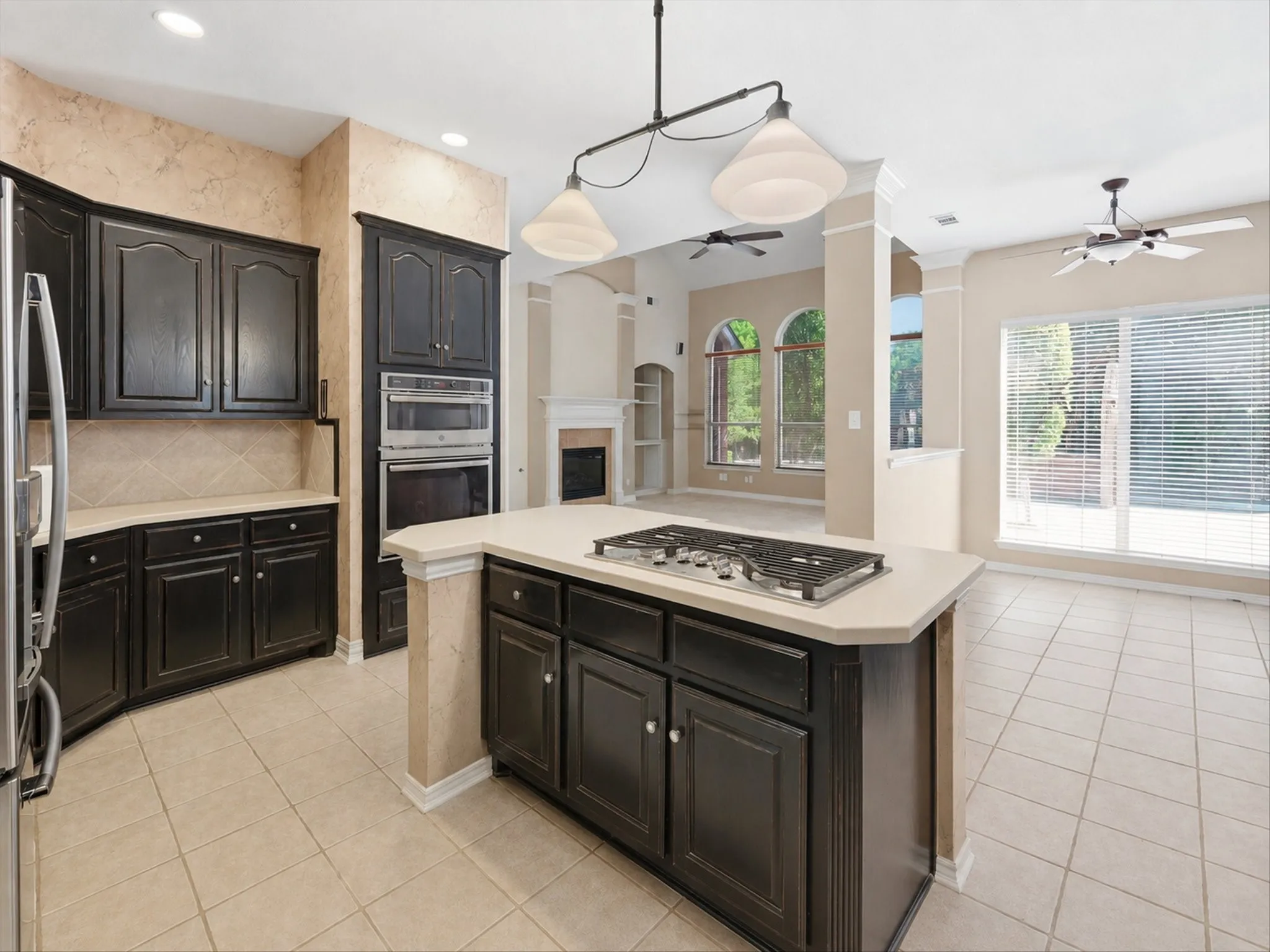 Open-concept kitchen featuring an island with a built-in gas cooktop, dark wood cabinetry, integrated stainless steel double ovens, tile flooring, and recessed lighting