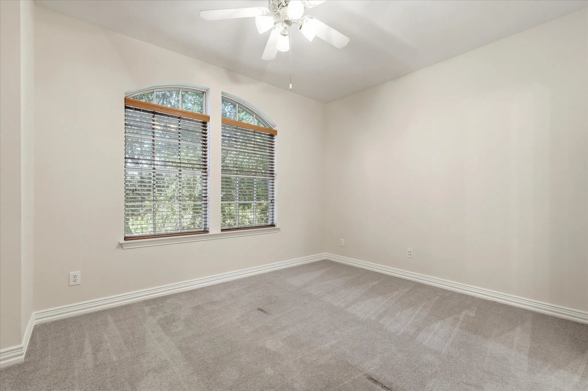 Bedroom featuring two arched windows with blinds, light beige wall paint, gray carpet flooring, white baseboards, and a ceiling fan
