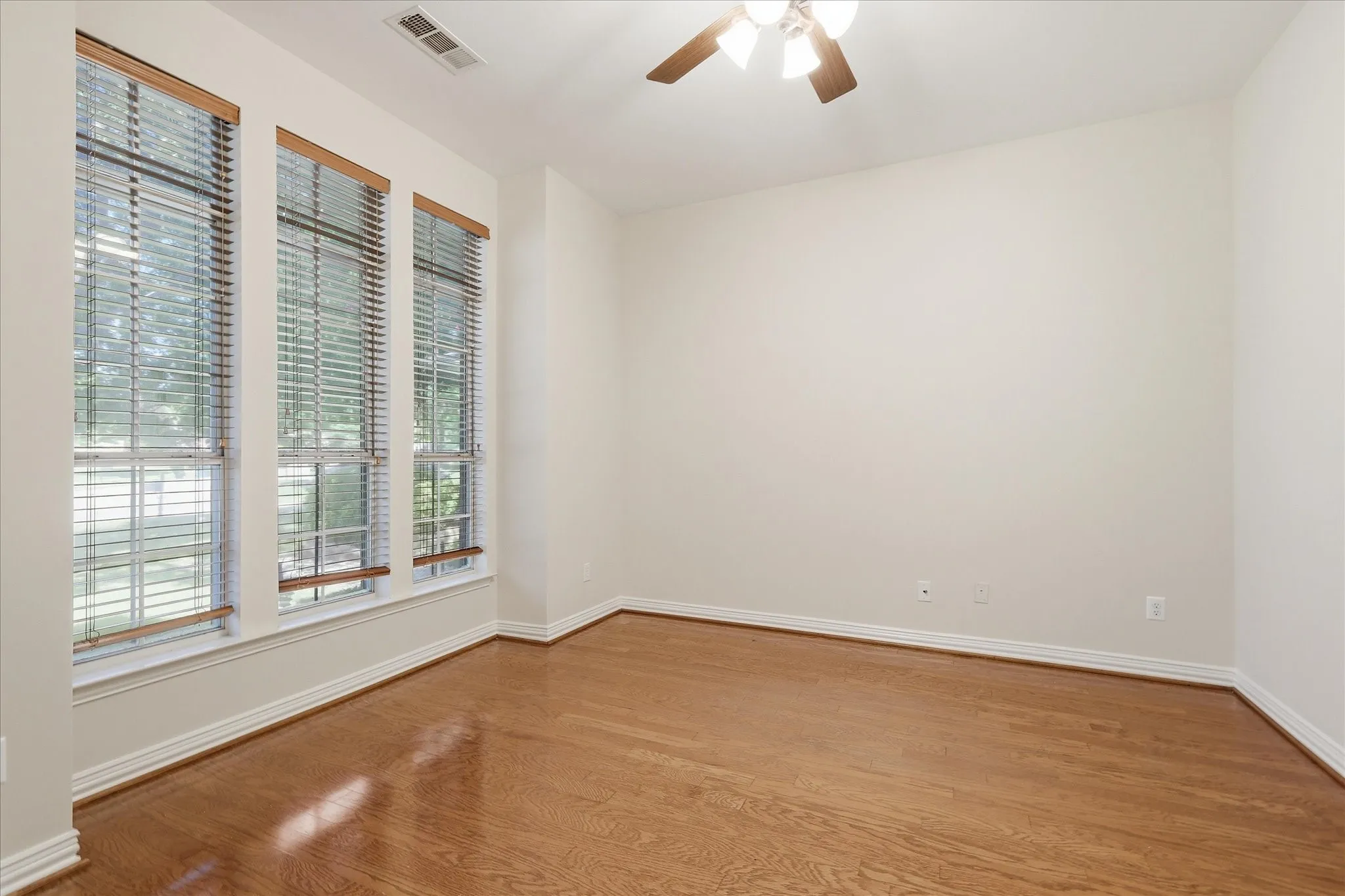 Room featuring wood-finish flooring, three large windows with blinds, a ceiling fan with light fixture, and white baseboards