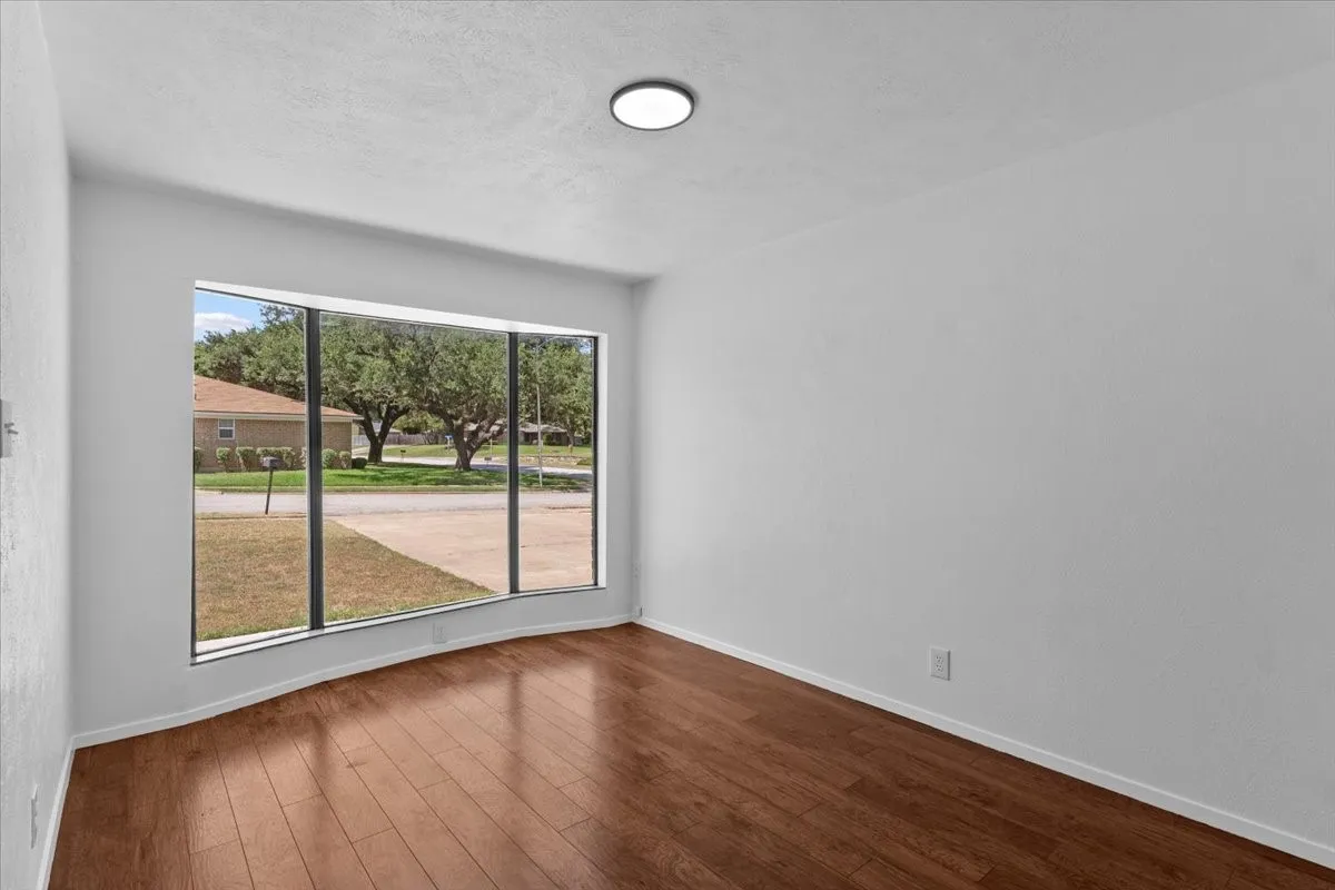 Spacious room featuring a large curved bay window, rich wood-finish flooring, bright white walls, and a recessed ceiling light fixture