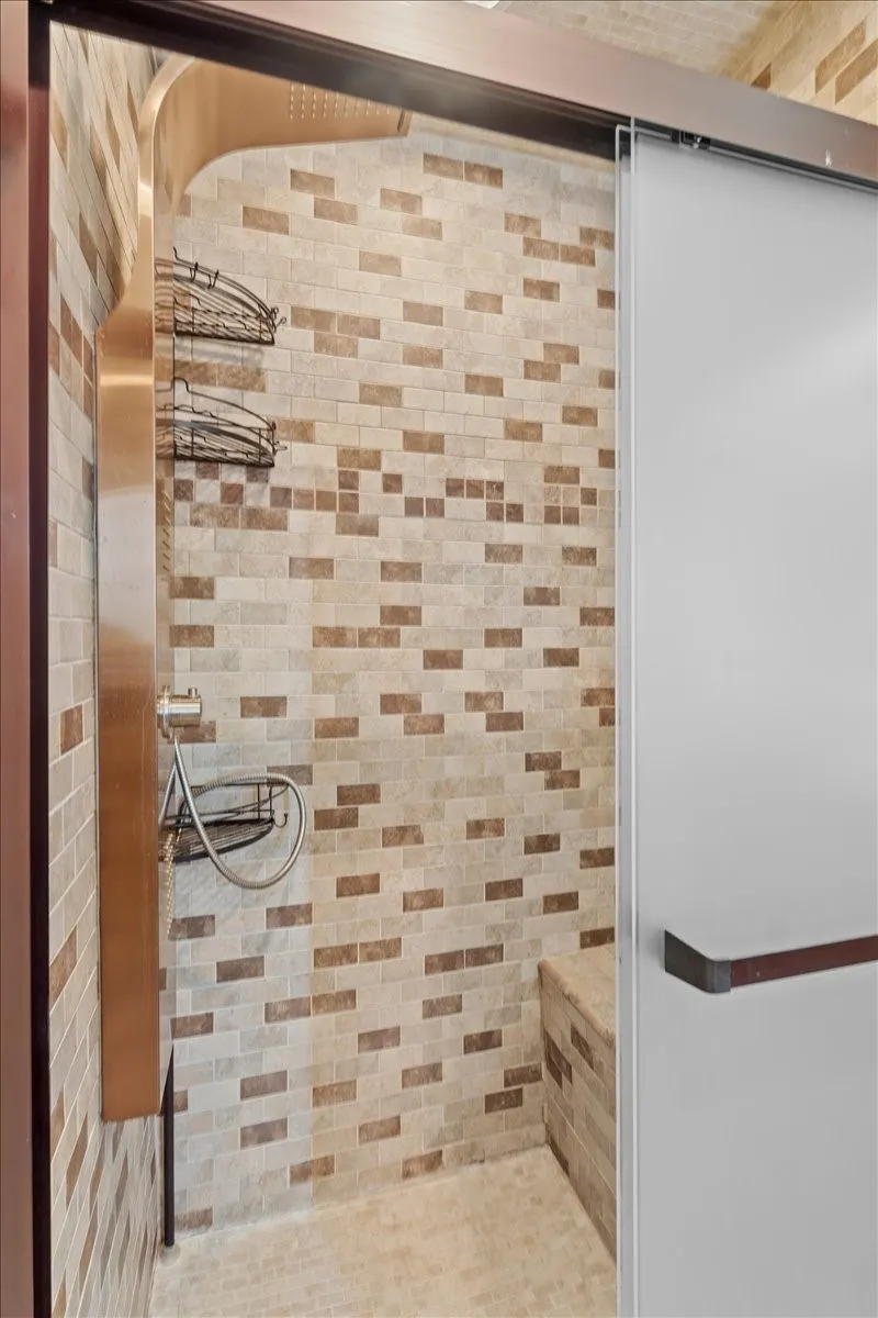 Enclosed shower featuring a textured tiled wall, built-in bench, and a brushed metal shower system with a handheld spray