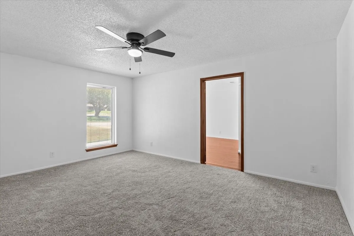 Spacious room featuring light gray carpeting, crisp white walls, a multi-pane window with wood-finish trim, and a ceiling fan with integrated lighting
