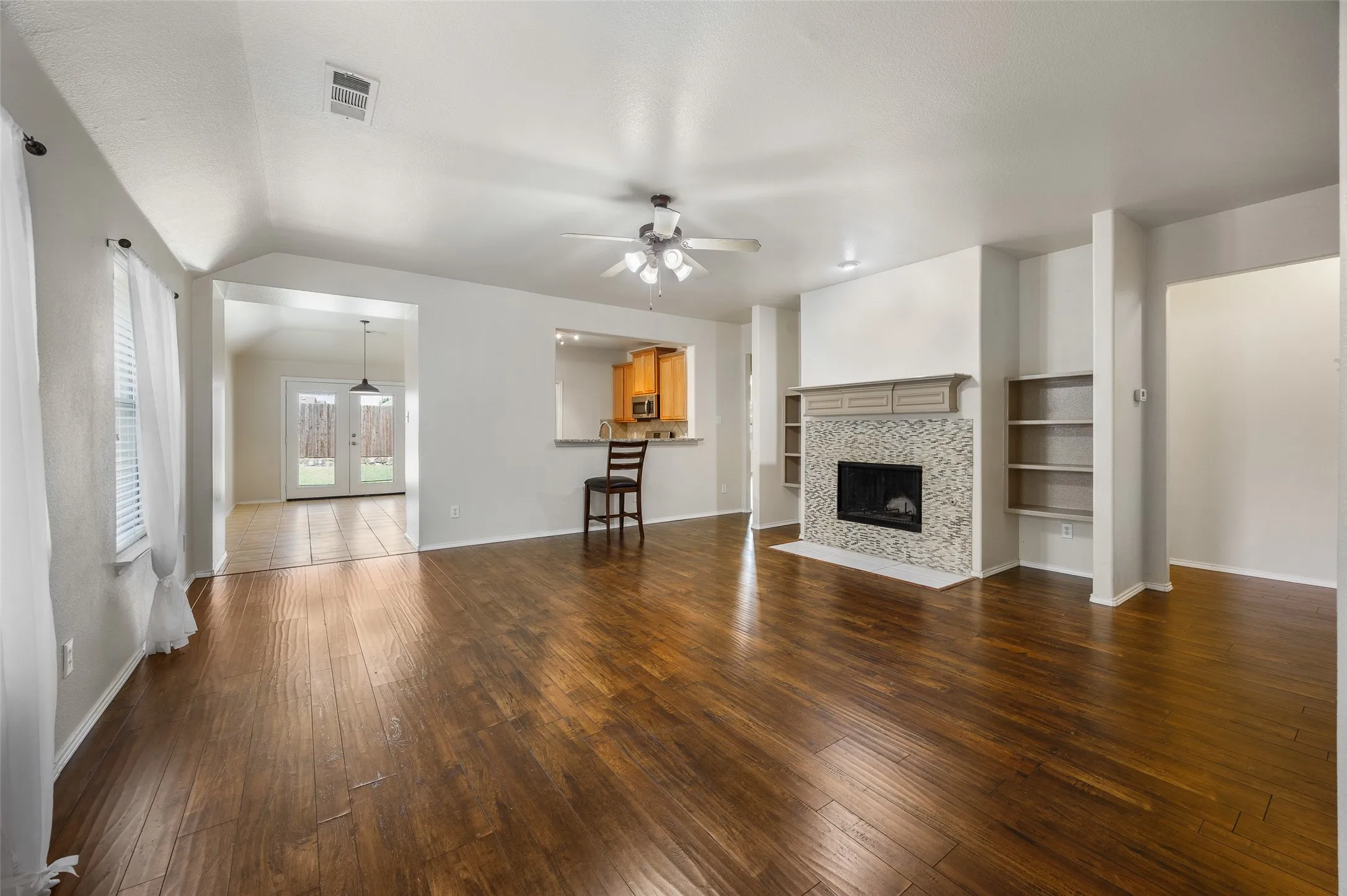 Spacious living area featuring wood-finish flooring, a tiled fireplace with mantel, built-in shelving, and a ceiling fan