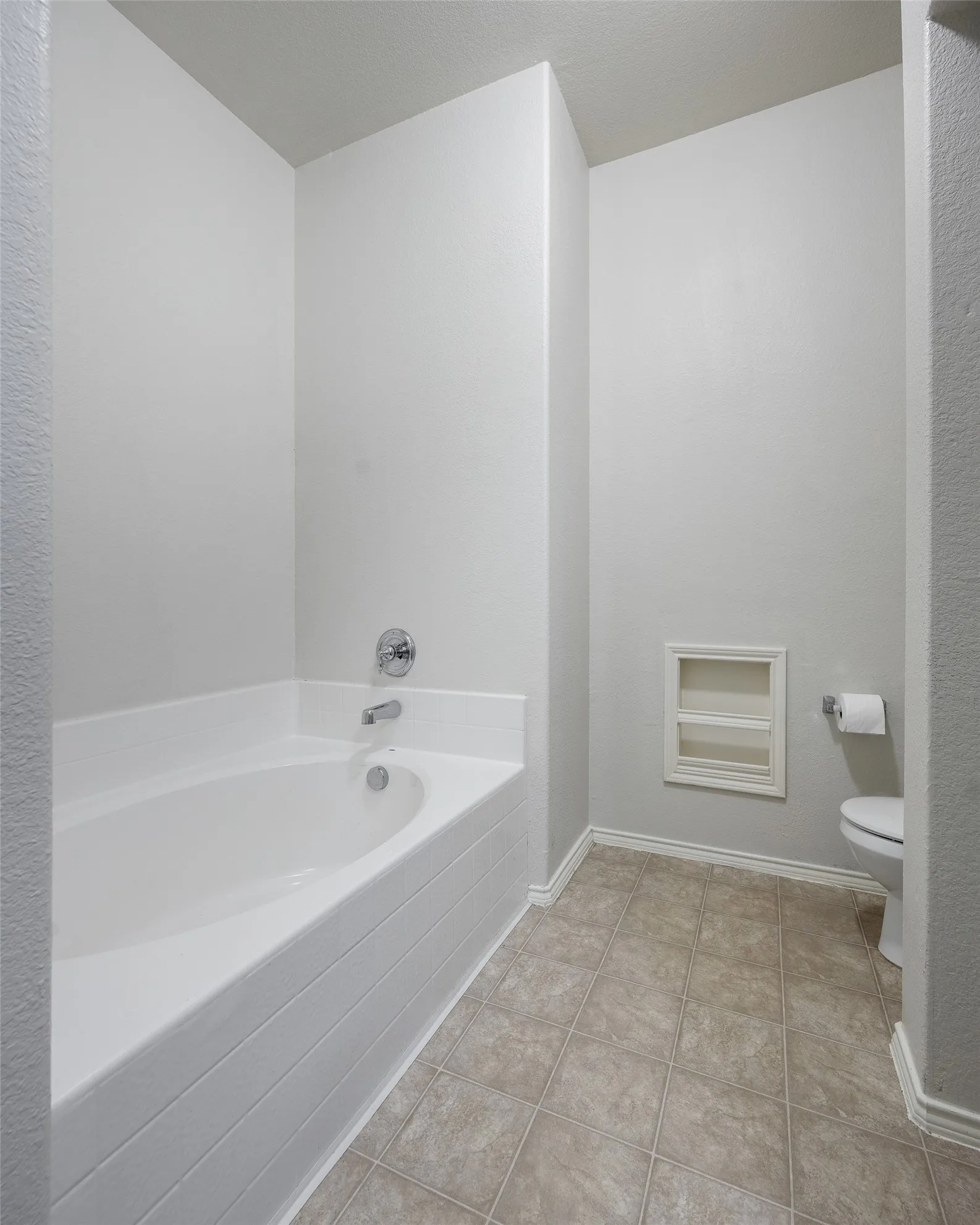 Bathroom with a built-in tub featuring chrome fixtures, textured tile flooring, and an integrated wall shelf