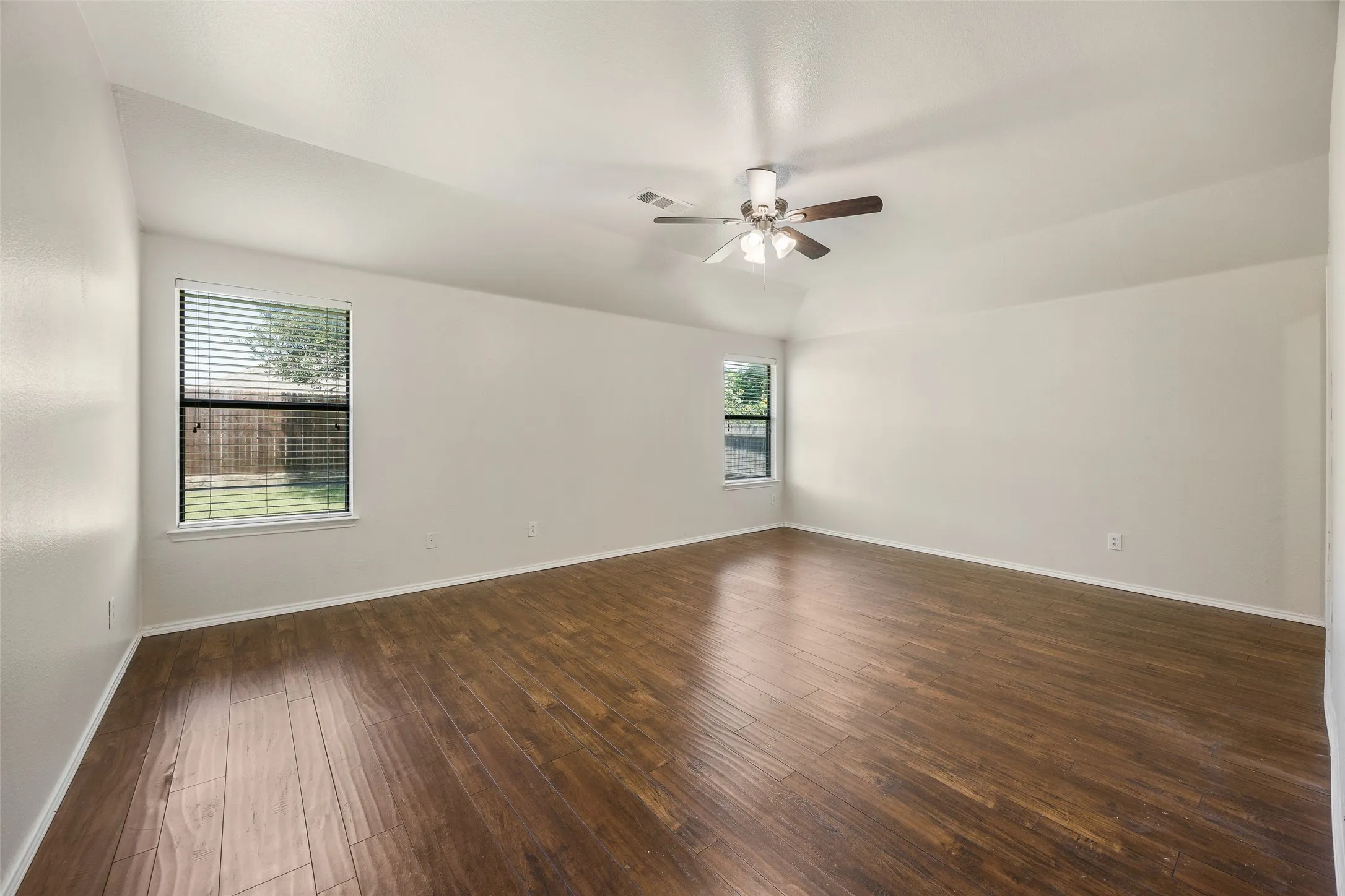 Spacious room featuring vaulted ceilings, wood-finish flooring, two windows with blinds, and a ceiling fan with integrated lighting