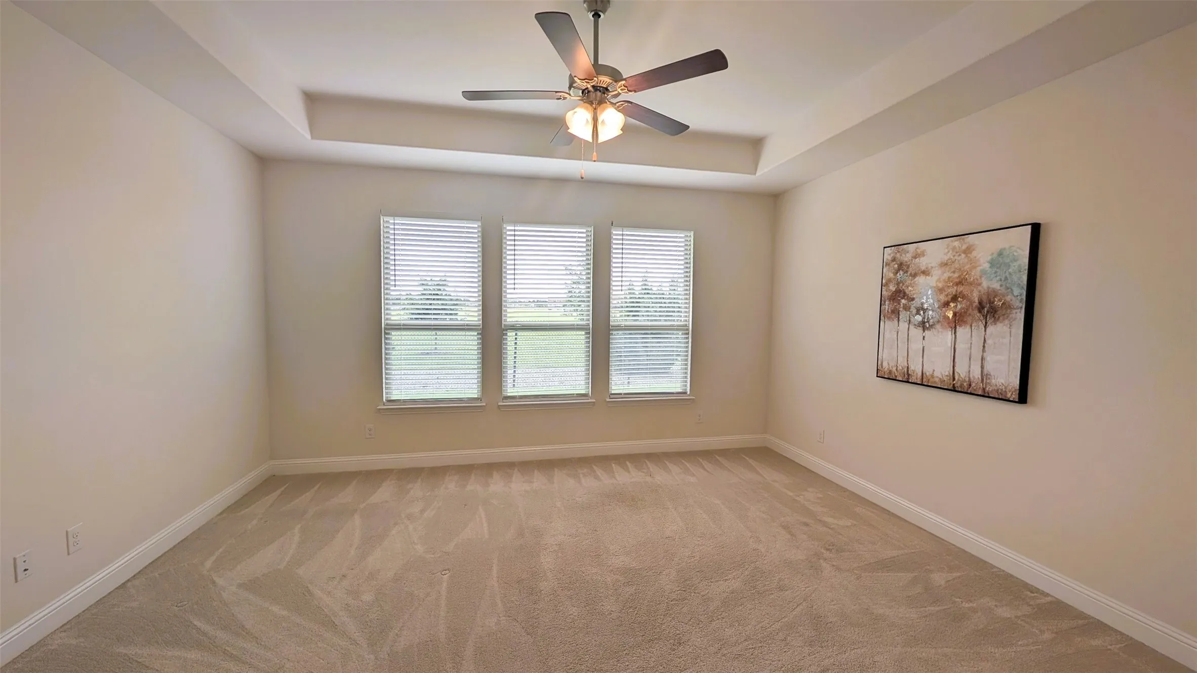 Spacious room featuring a tray ceiling, ceiling fan with light fixture, three windows with horizontal blinds, and neutral carpet flooring