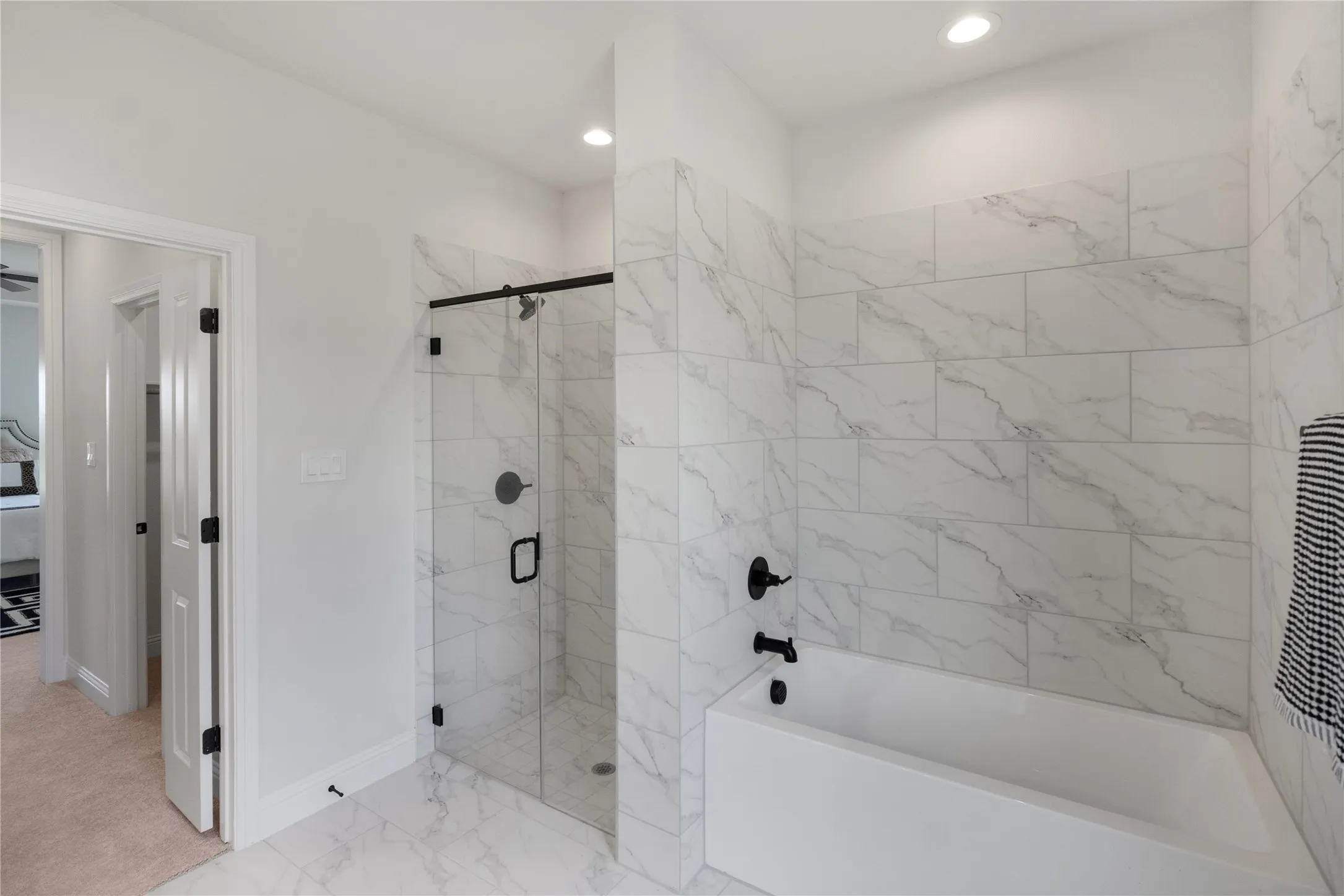 Full bathroom featuring a white soaking tub with black fixtures, a separate glass-enclosed shower with black hardware, and white marble-look tile flooring and shower​​‌​​​​‌​​‌‌​‌‌​​​‌‌​‌​‌​‌​​​‌​​ surround