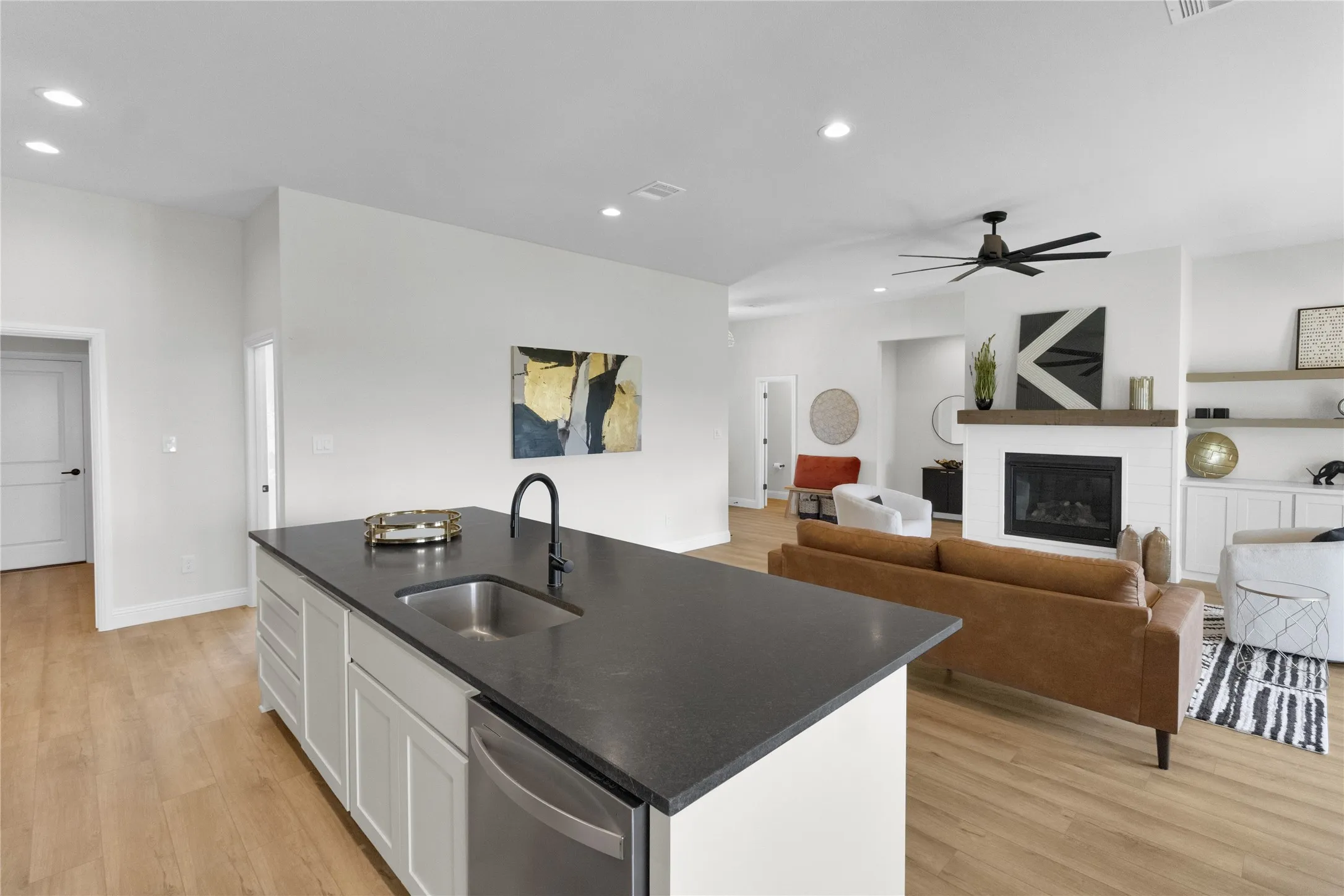 Open-concept living area featuring a kitchen island with dark countertop and stainless steel sink, wood-finish flooring, recessed lighting, and a fireplace with a wood mantle