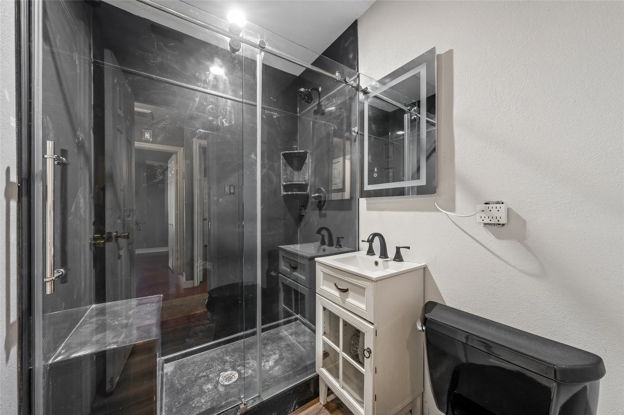 Bathroom featuring a glass-enclosed shower with black-toned wall panels, a white vanity with integrated sink, a black toilet, and an illuminated medicine cabinet