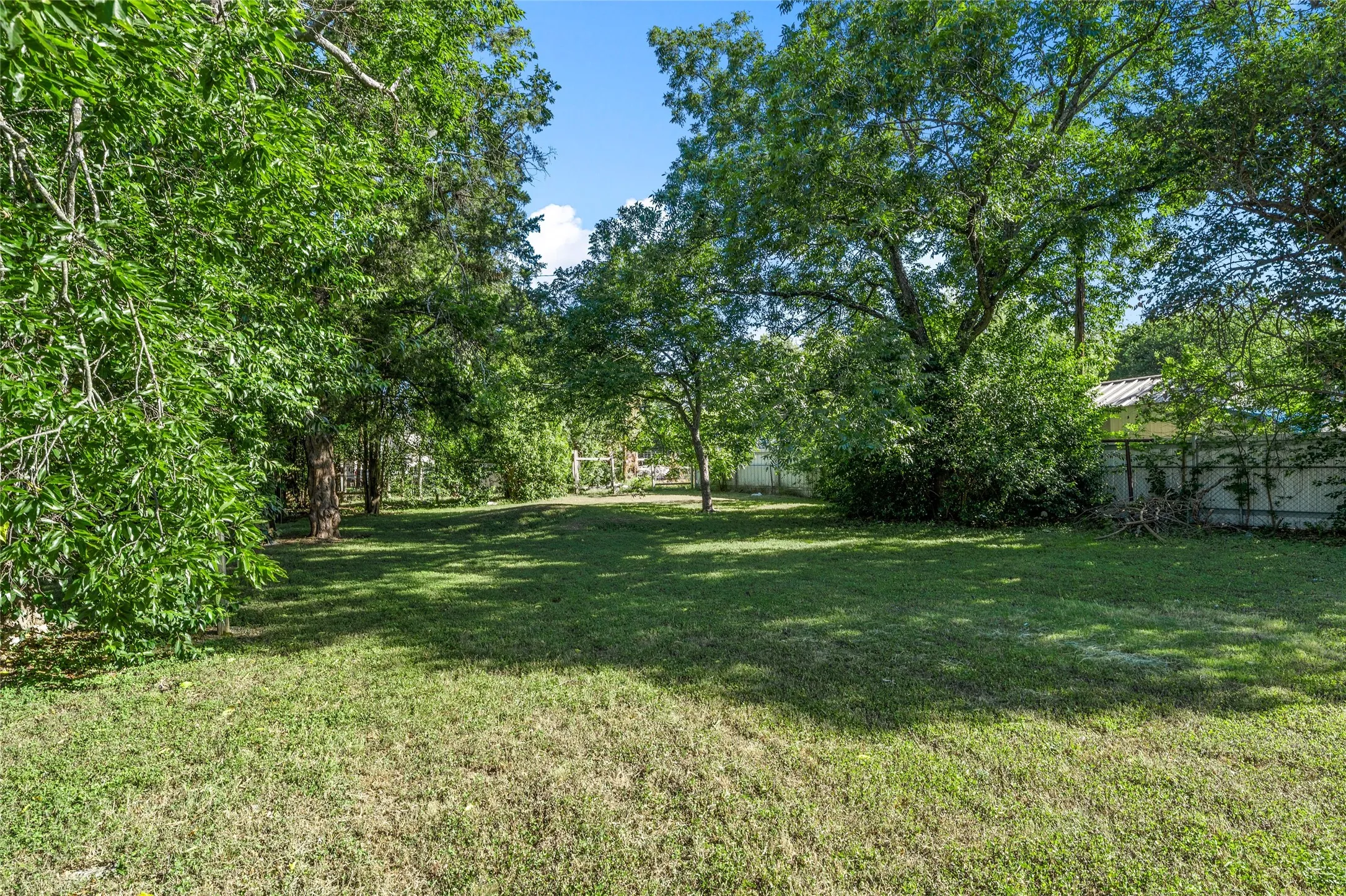 Expansive grassy lawn surrounded by mature trees, featuring a tree-lined perimeter and a partial privacy fence