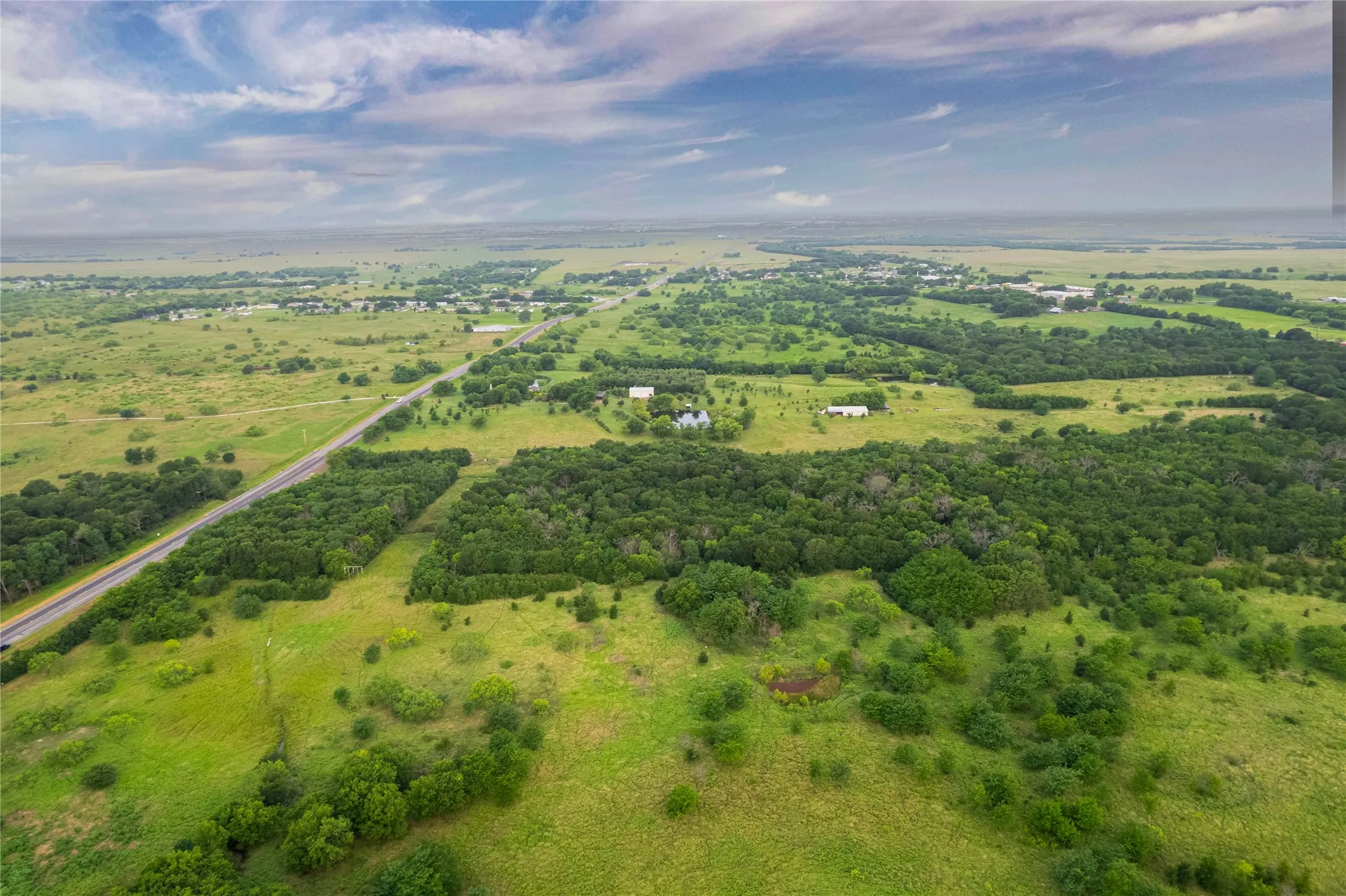 Expansive land parcel featuring a main road, scattered buildings, dense tree coverage, open grassy areas, and a visible pond