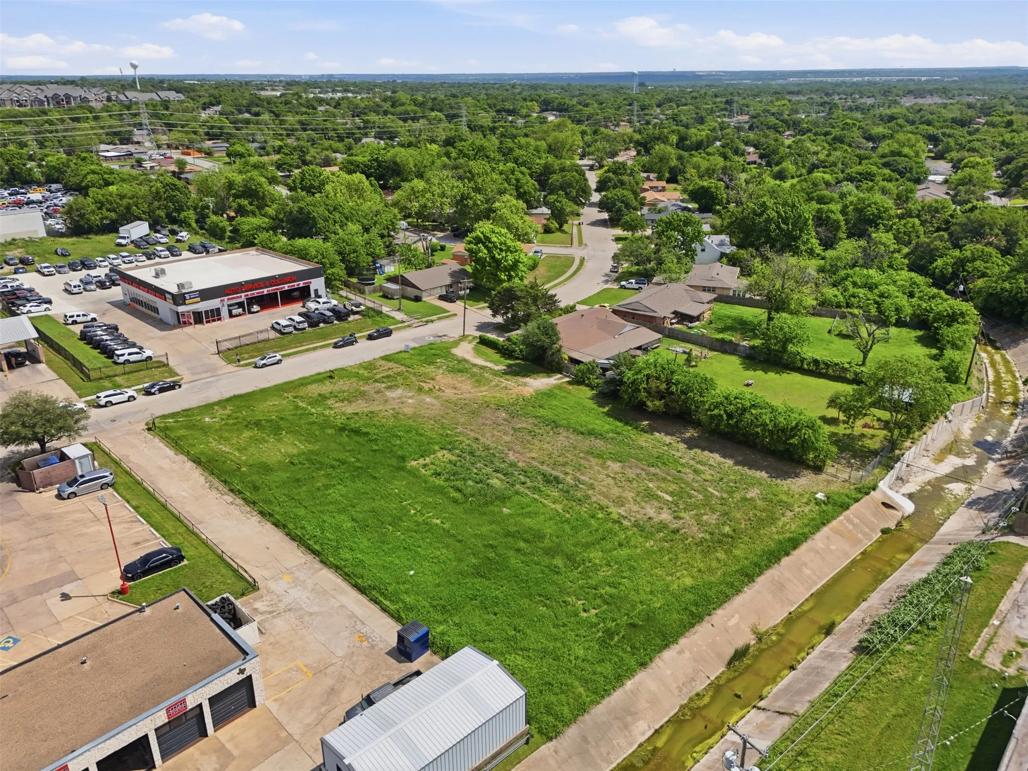 Expansive vacant lot featuring a grass expanse, concrete perimeter, and established tree coverage in the surrounding area