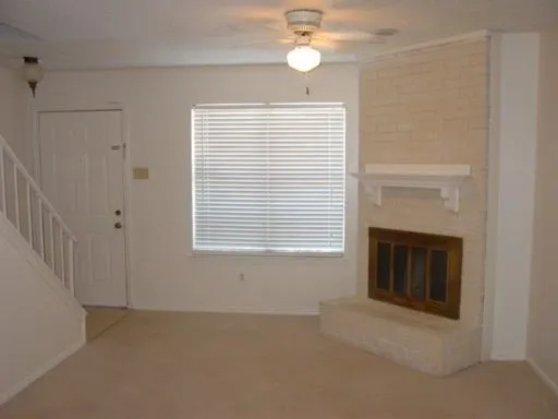 Living area featuring a painted brick fireplace with a wood-finish insert, an overhead ceiling fan with lighting, a window with horizontal blinds, and carpeted flooring