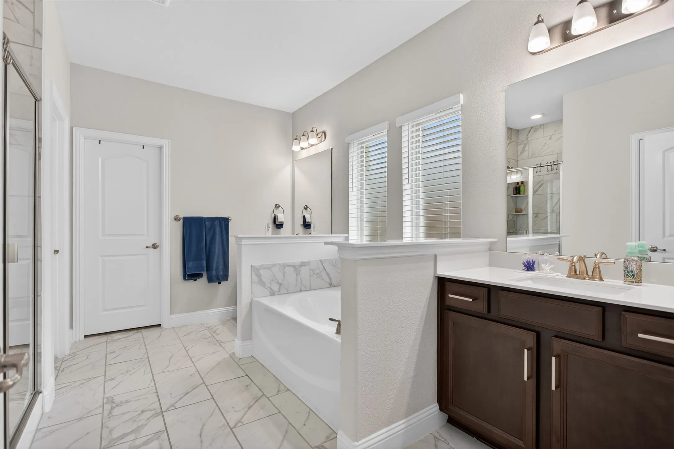 Spacious bathroom featuring a built-in soaking tub with marble-patterned tile surround, dual windows with horizontal blinds, and a double vanity with dark wood cabinetry