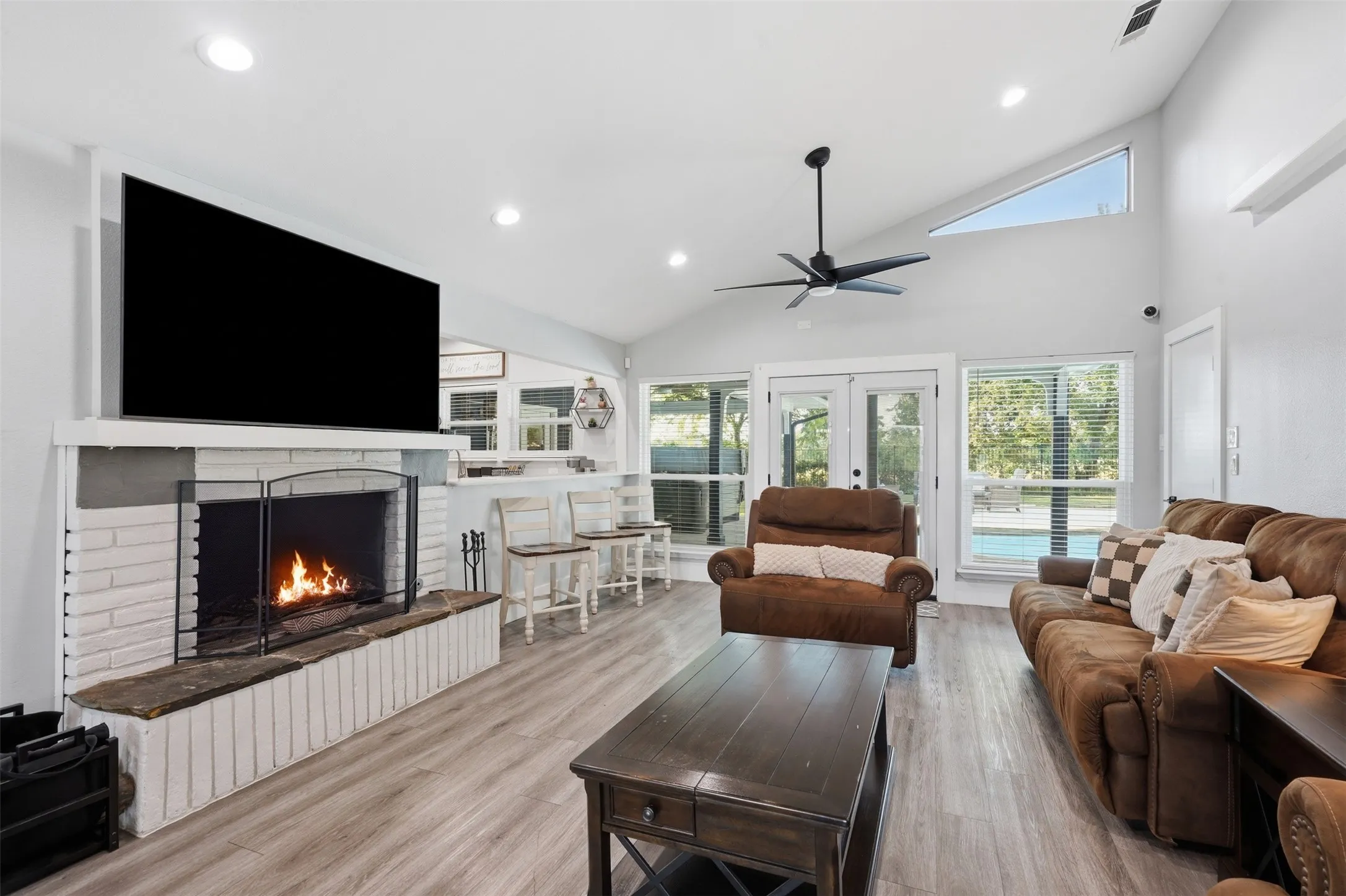 Spacious living area featuring a white painted brick fireplace with hearth, wood-finish flooring, vaulted ceilings, and a modern ceiling fan