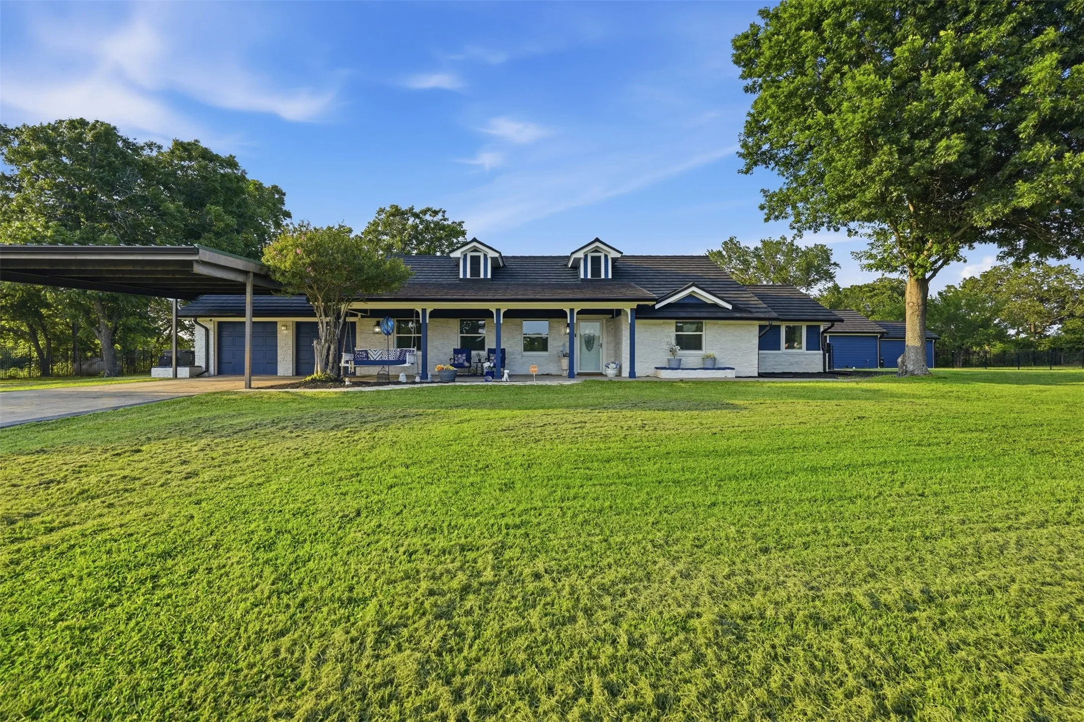 Expansive front lawn with mature trees frames a white brick home featuring a covered front porch, dormer windows, and an attached carport