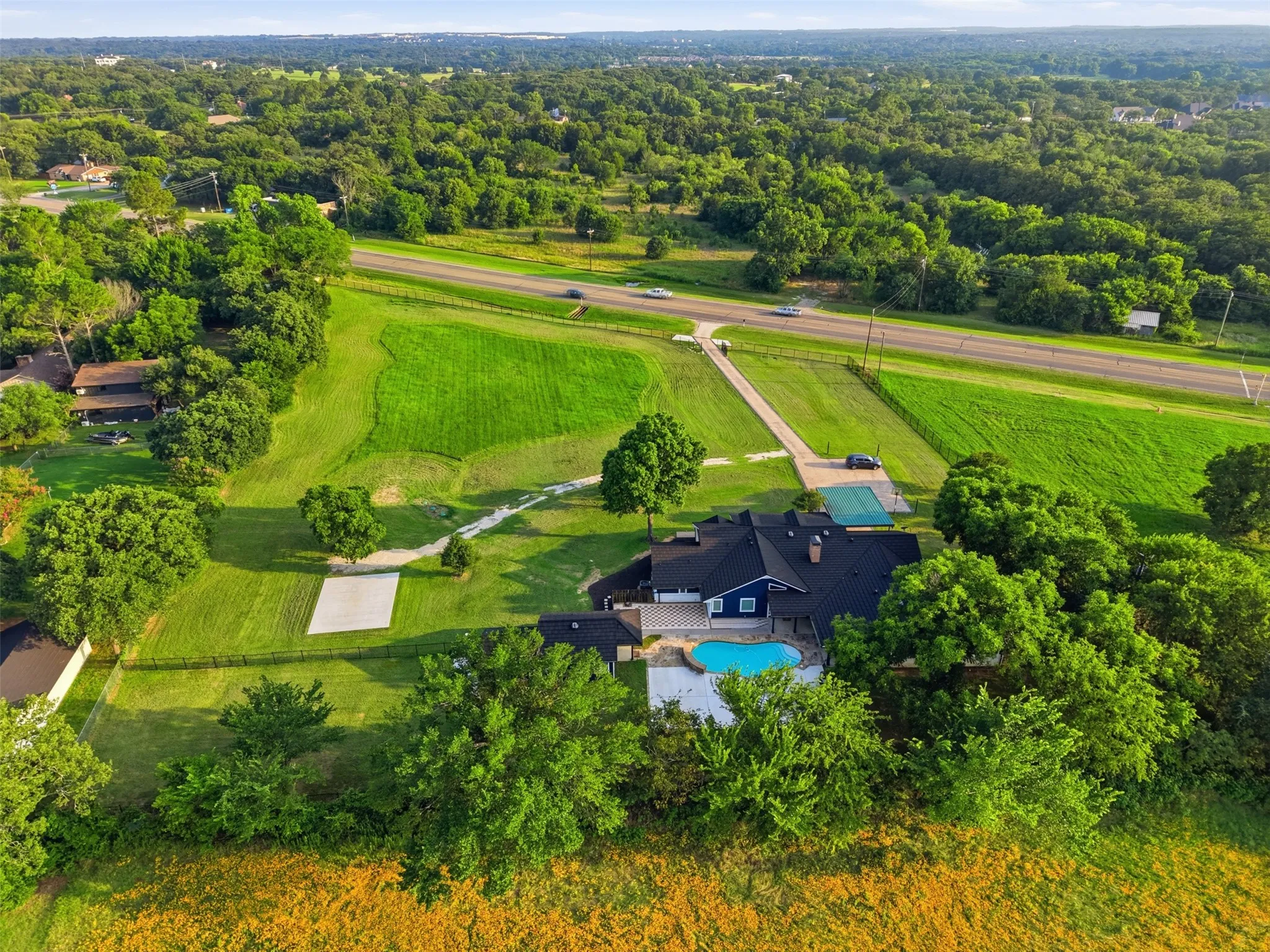 Aerial property view featuring a dark-hued residence with a backyard swimming pool, expansive green lawns, mature trees, and a visible roadway