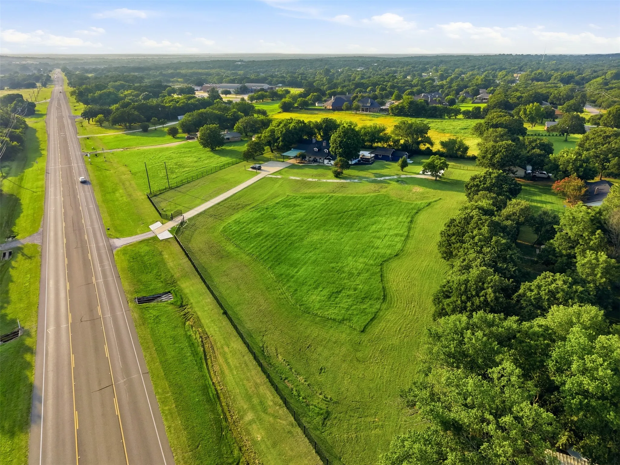 Expansive green landscape with a paved road running alongside, featuring wooded borders and a distant residential area