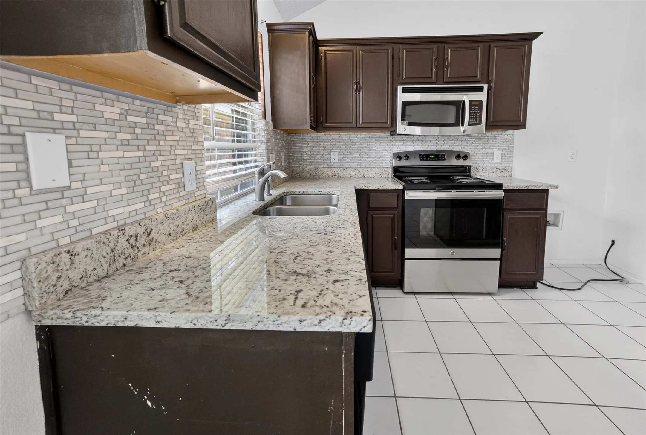 Kitchen featuring granite countertops, a mosaic tile backsplash, a double basin stainless steel sink, stainless steel appliances, and dark wood cabinetry