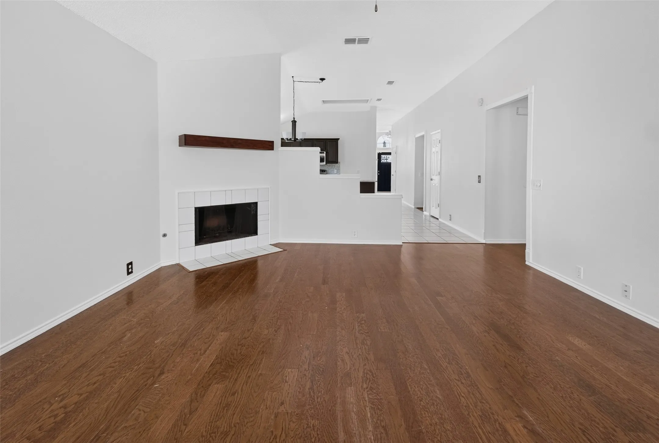 Spacious living area featuring wood-finish flooring, a white tiled fireplace with a dark wood mantel, and crisp white walls