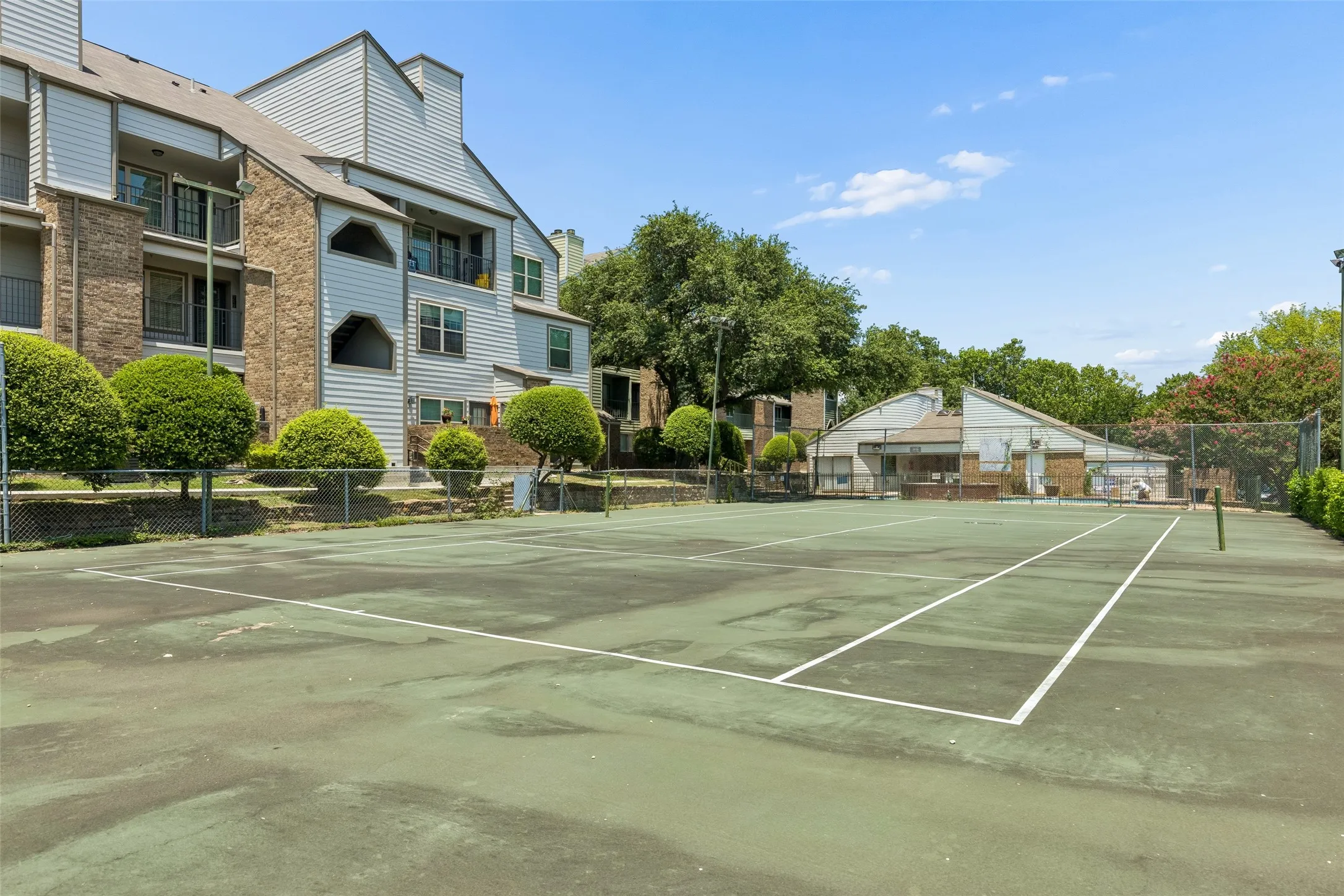 Outdoor tennis court with perimeter fencing, surrounded by mature trees and landscaping