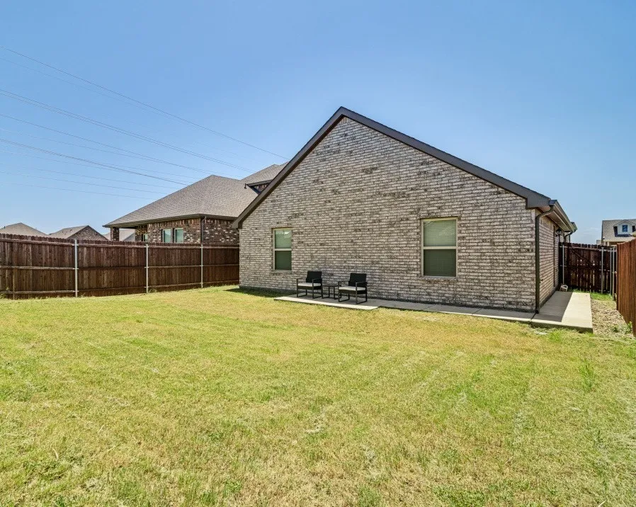 Expansive backyard featuring a manicured lawn, full privacy fencing, a concrete patio, and a brick exterior with dark trim