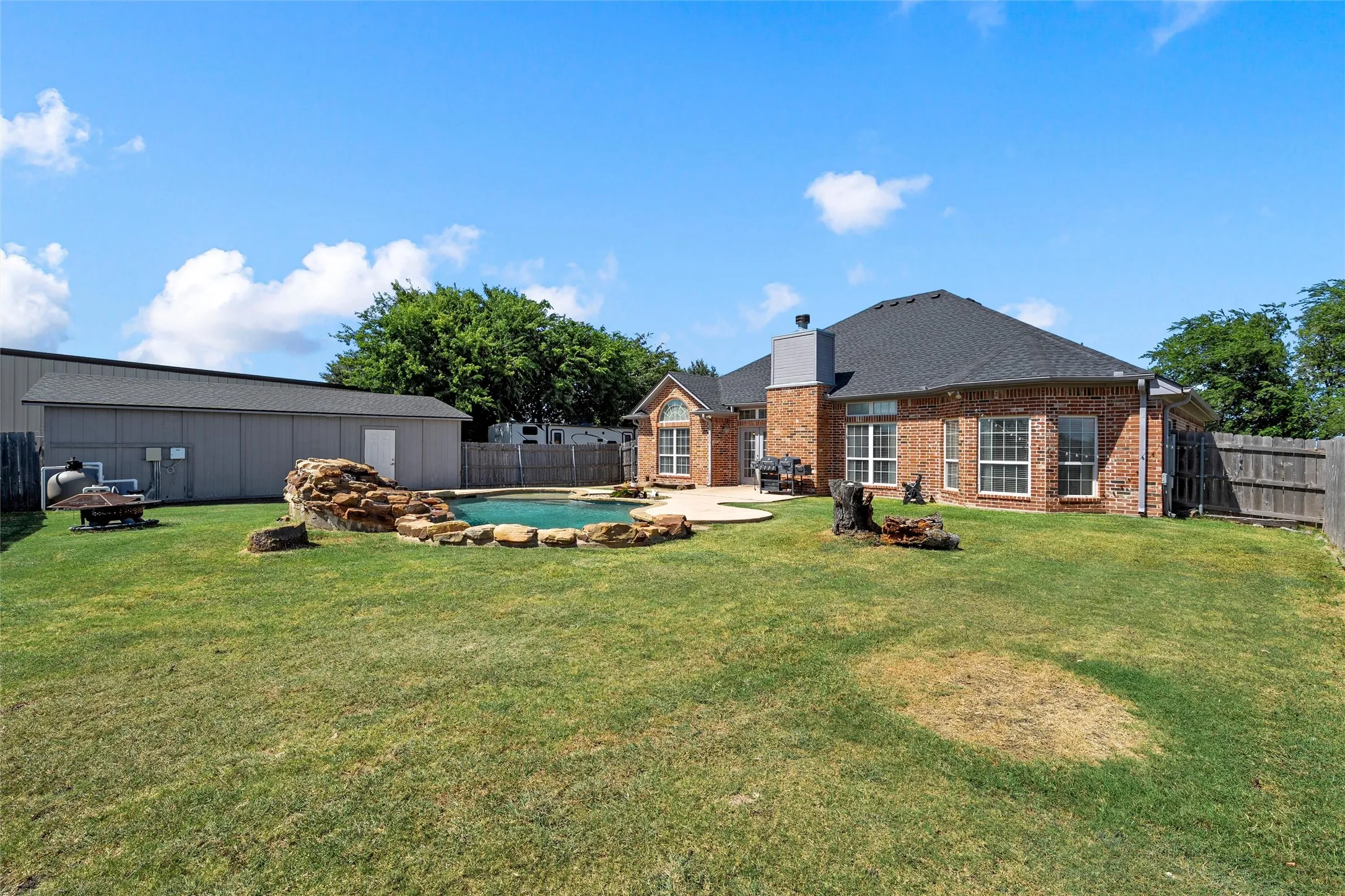 Expansive backyard featuring a sparkling in-ground pool with rock detailing, a concrete patio, and a large detached outbuilding