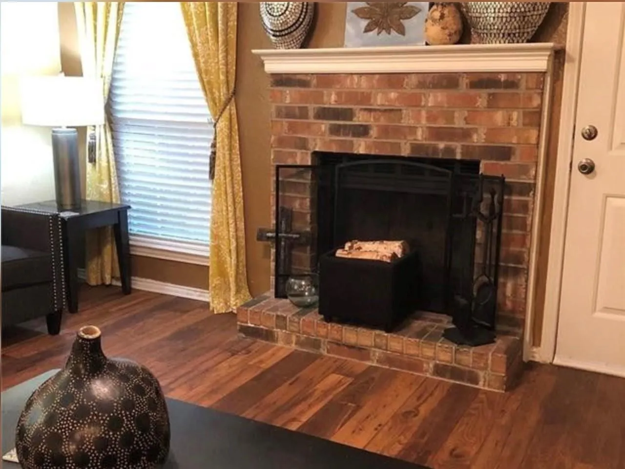 Brick fireplace with a white mantel, complemented by wood-finish flooring, a window with blinds, and a white paneled door