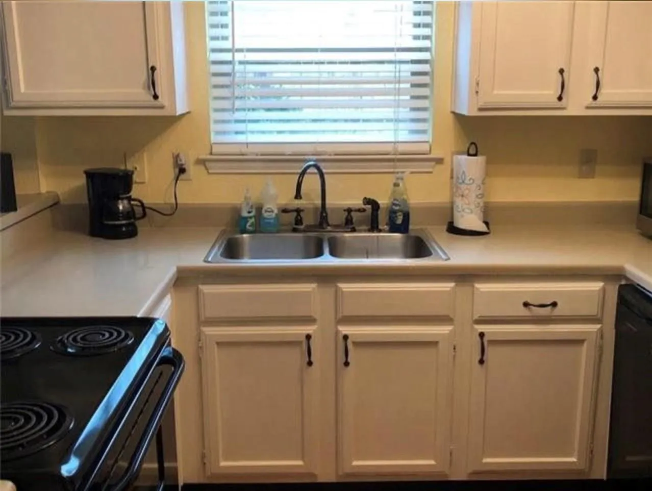 Kitchen sink area featuring a dual basin stainless steel sink, black faucet, white shaker-style cabinetry with dark hardware, and light-toned countertops