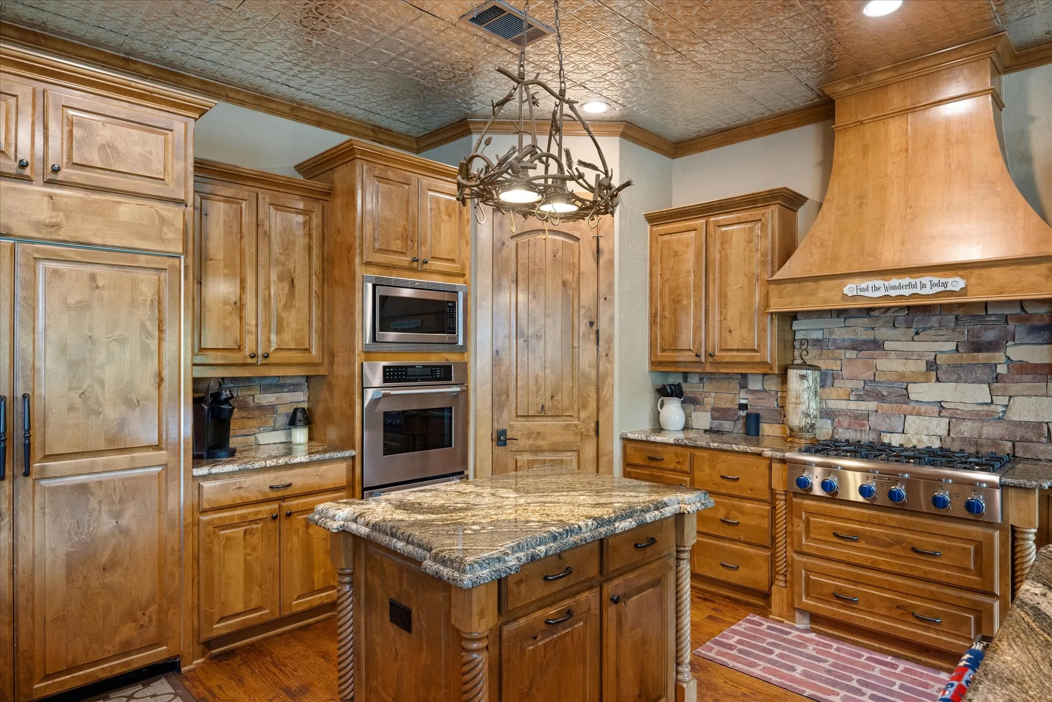 Kitchen featuring custom wood cabinetry, stainless steel appliances, a stone backsplash, granite countertops, and a decorative pressed tin ceiling